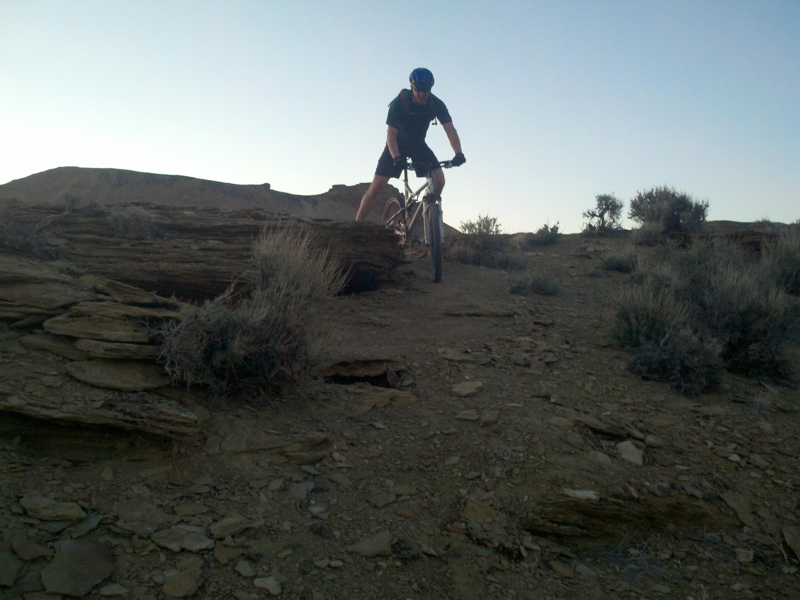 A mountain biker navigating a rocky terrain during sunset, with shrubs and desert landscape in the background. MacBones Trail mountain bike trail.