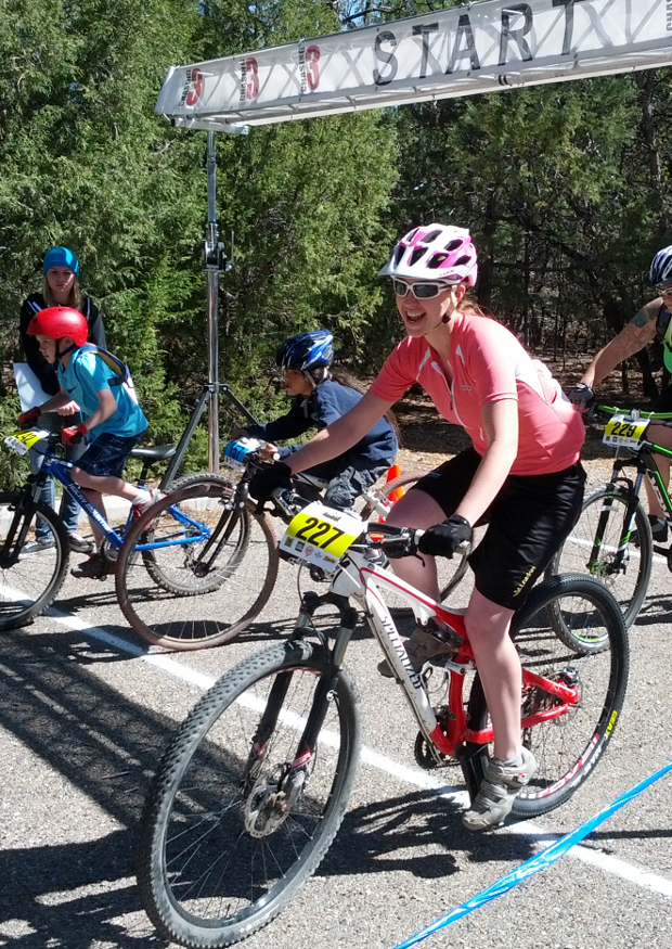 A young woman wearing a pink shirt and helmet smiles while riding a mountain bike at the starting line of a cycling event. Several other participants are visible in the background, and a "START" banner can be seen overhead. The scene is set outdoors, surrounded by greenery.