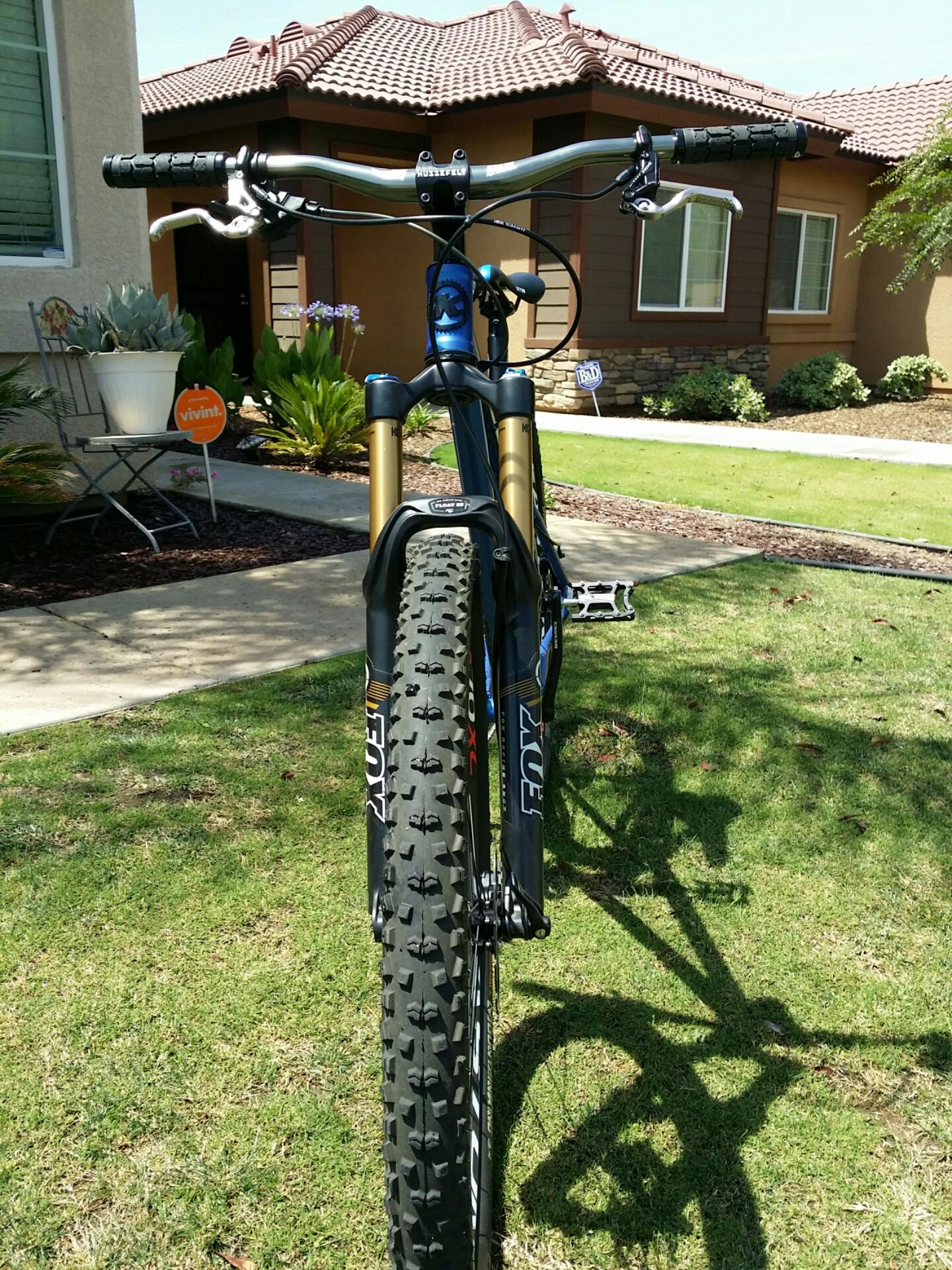Kona Honzo: A close-up view of a mountain bike from the front, featuring a fork with black and gold components, thick knobby tires, and handlebars with grips. The bike is positioned on a grassy area, with a residential home and landscaping visible in the background, including plants and a small chair. Bright sunlight casts a shadow of the bike onto the grass.