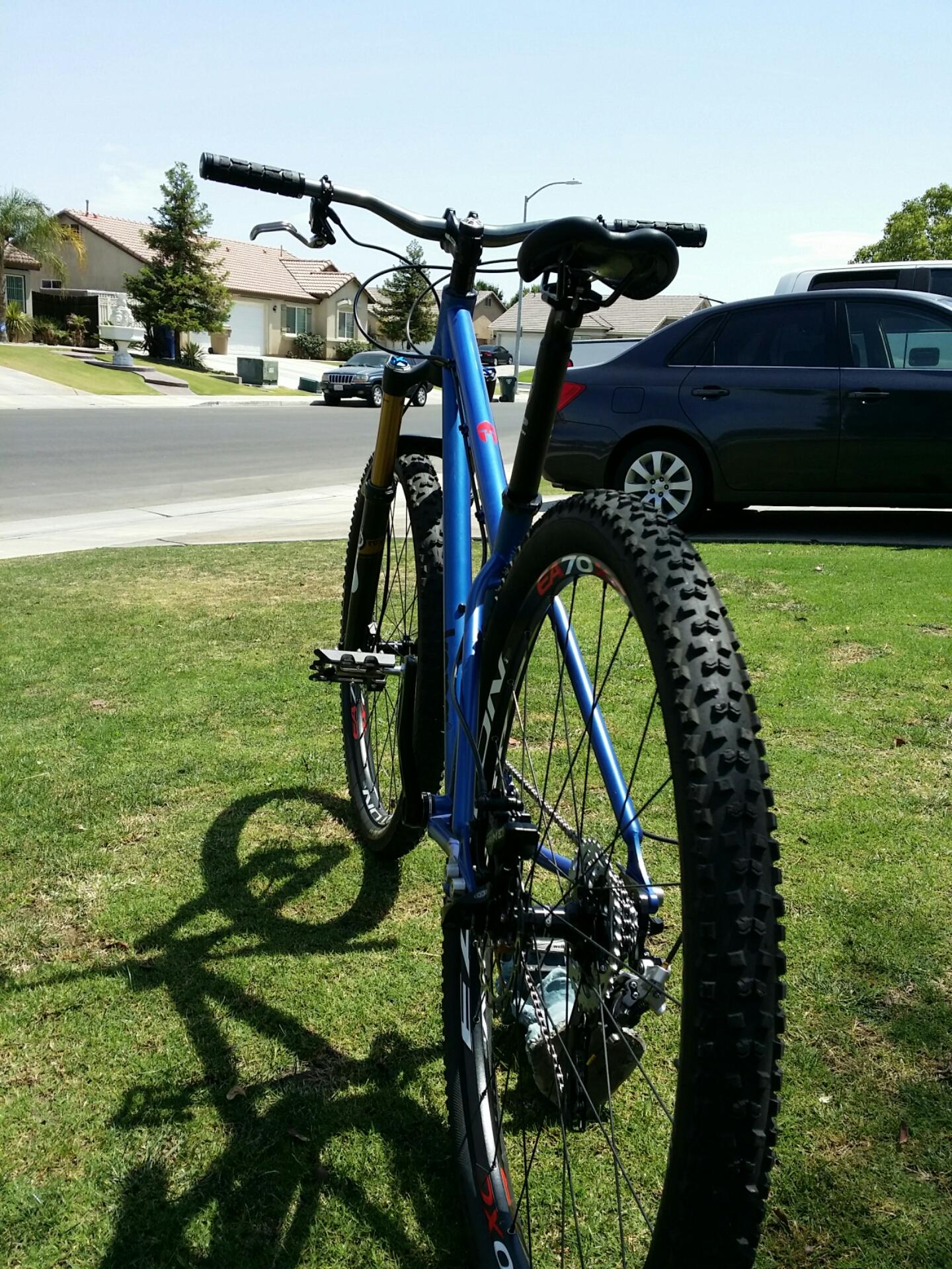 Kona Honzo: A blue mountain bike parked on grassy terrain, with the foreground showing the rear end and wheels. In the background, a suburban street is visible with houses and parked cars under a clear blue sky.