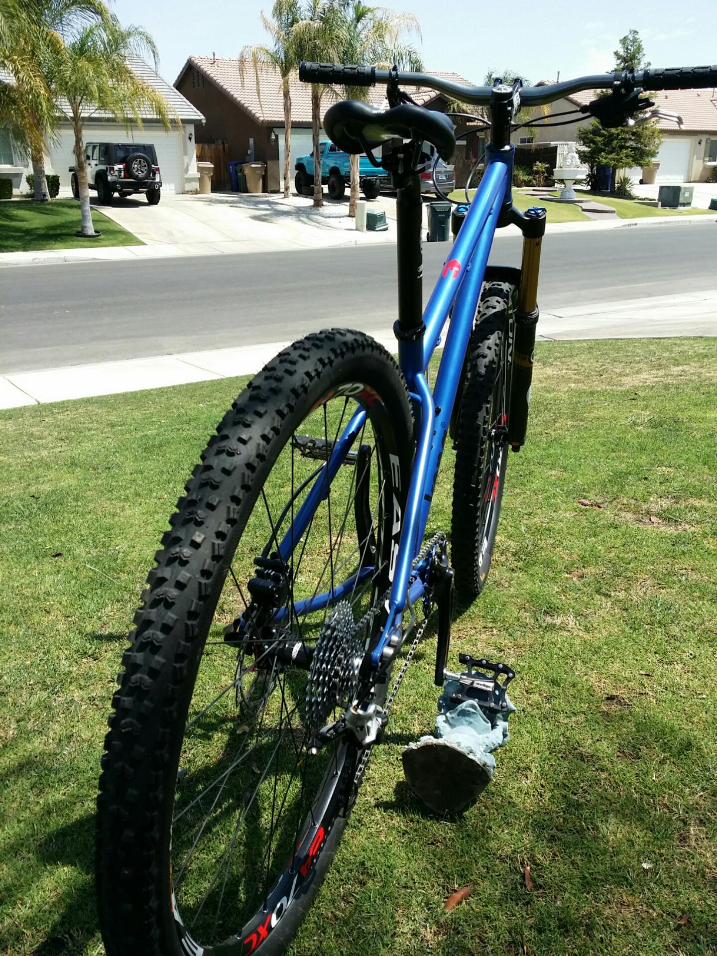 Kona Honzo: A blue mountain bike with thick tires is positioned on a grassy lawn, showcasing its rear. The bike features a modern design with a visible suspension system and pedal. In the background, suburban houses and parked vehicles can be seen under clear skies.