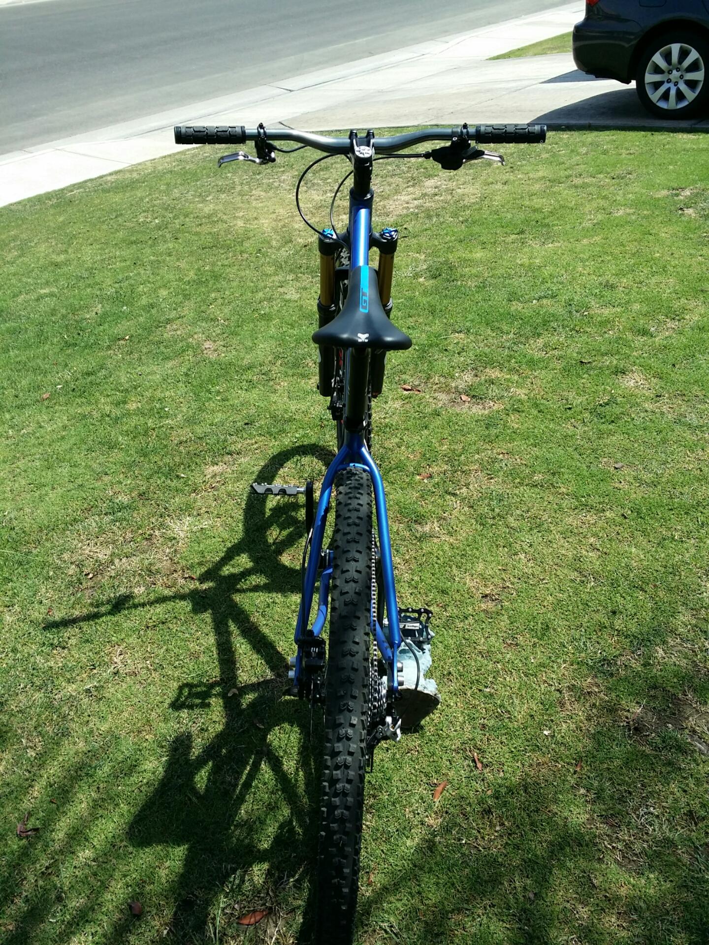 Kona Honzo: A blue mountain bike is positioned on a grassy area, viewed from the front. The bike features prominent front suspension and a sleek design, with handlebars and a black saddle clearly visible. In the background, there is a paved road and a parked car. The sunlight creates distinct shadows on the grass.
