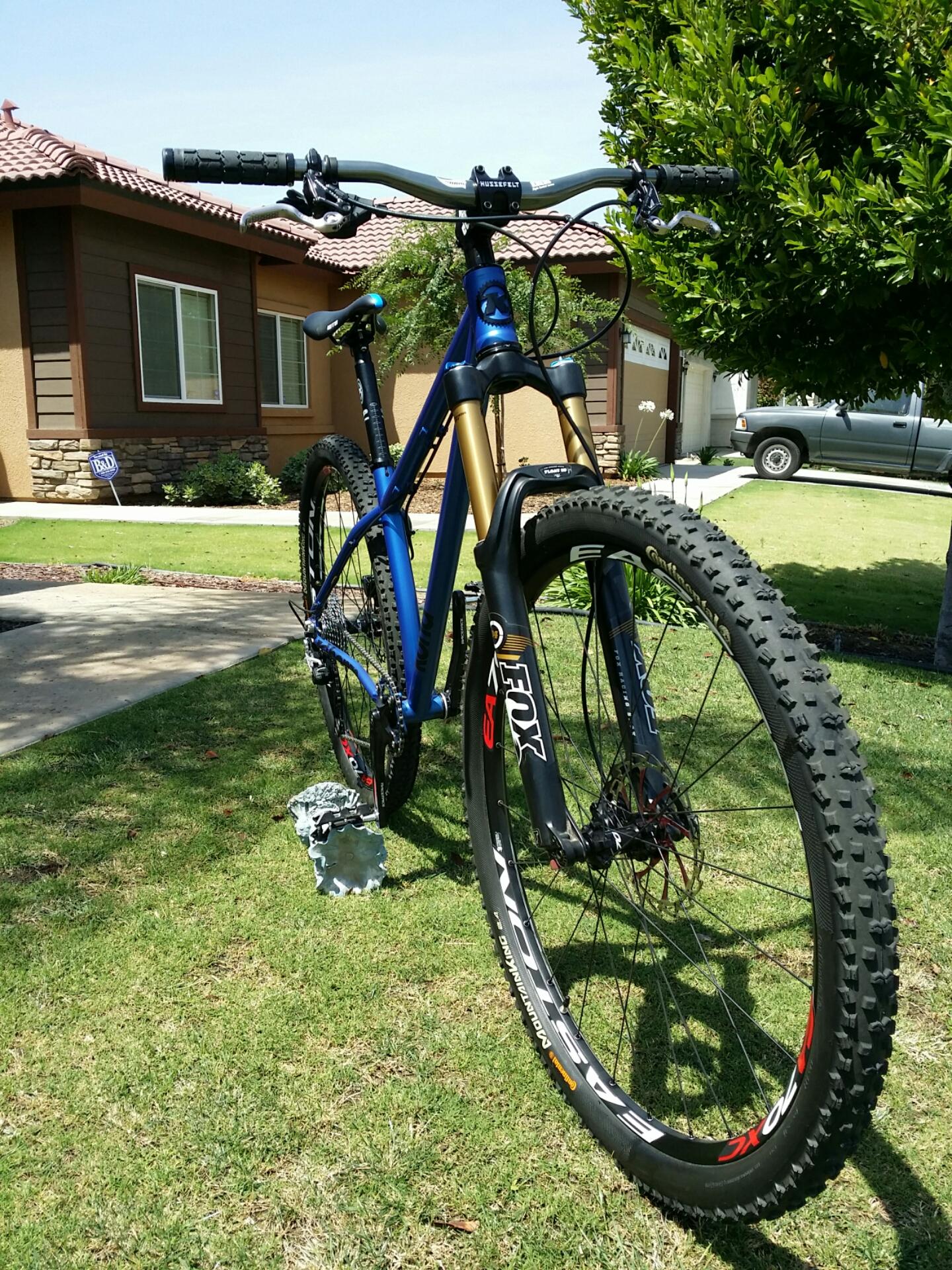 Kona Honzo: A mountain bike with a blue and gold frame is parked on a grassy lawn in front of a house. The bike features thick tires and a front suspension fork. In the background, a single-story home can be seen with a driveway and a gray pickup truck. A small decorative item is placed near the bike on the grass.
