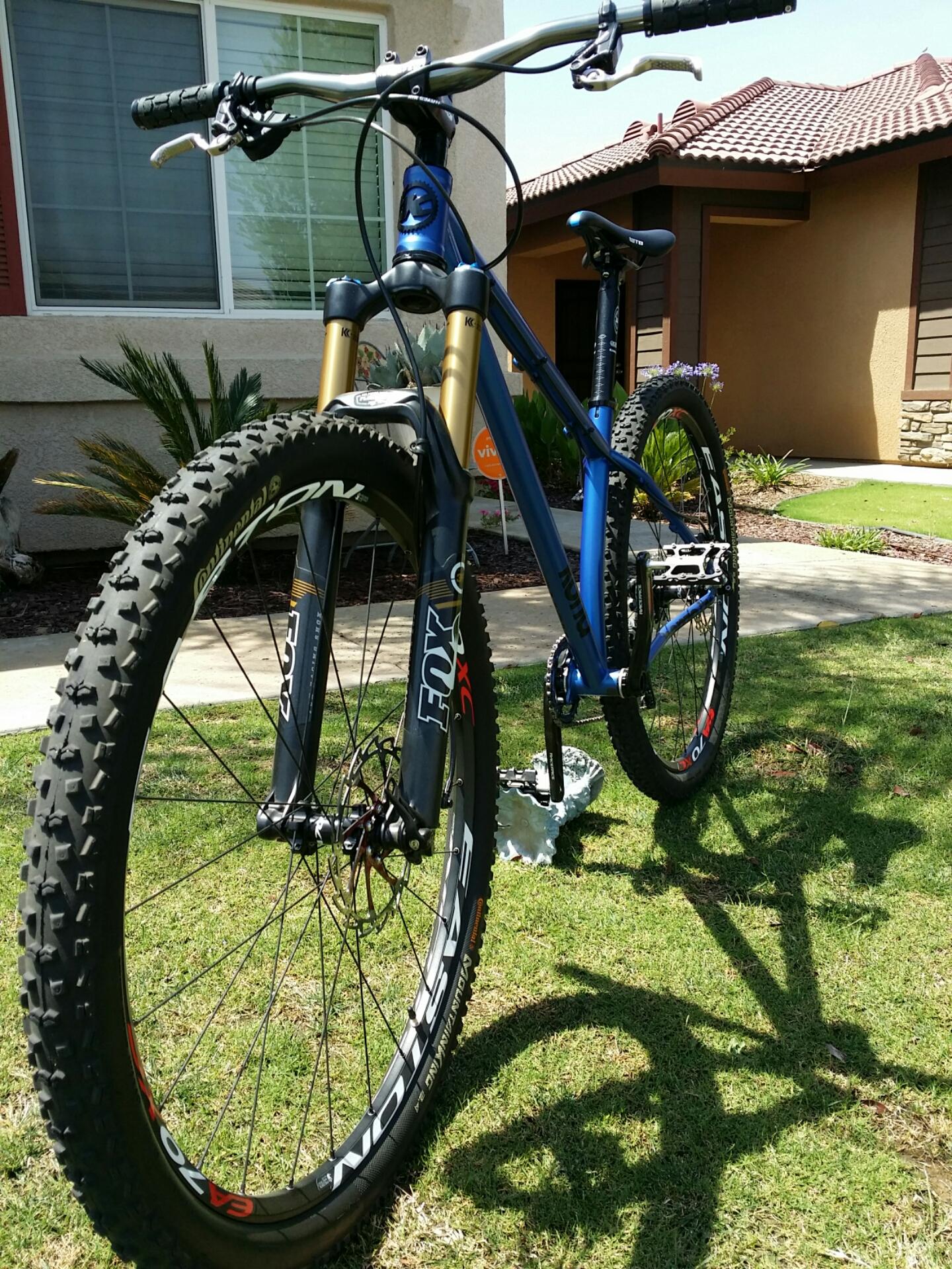 Kona Honzo: A blue mountain bike with gold suspension forks is parked on a grassy area in front of a house. The bike features wide, knobby tires designed for off-road terrain and has a sleek, modern design. In the background, there are residential elements like a well-maintained lawn, decorative plants, and a house with a tile roof. The scene is bright and sunny, highlighting the bike and its surroundings.