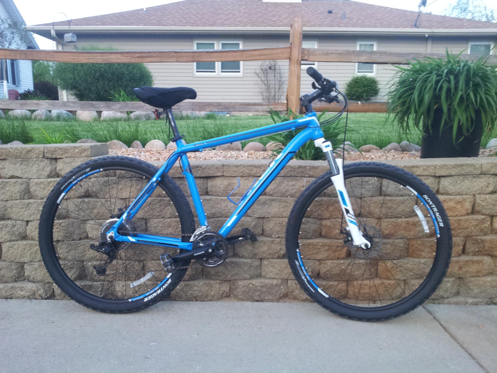 Trek Marlin: A blue mountain bike parked on a concrete surface next to a stone wall, with greenery and a house in the background. The bike features thick tires and a front suspension fork, reflecting its design for off-road riding.