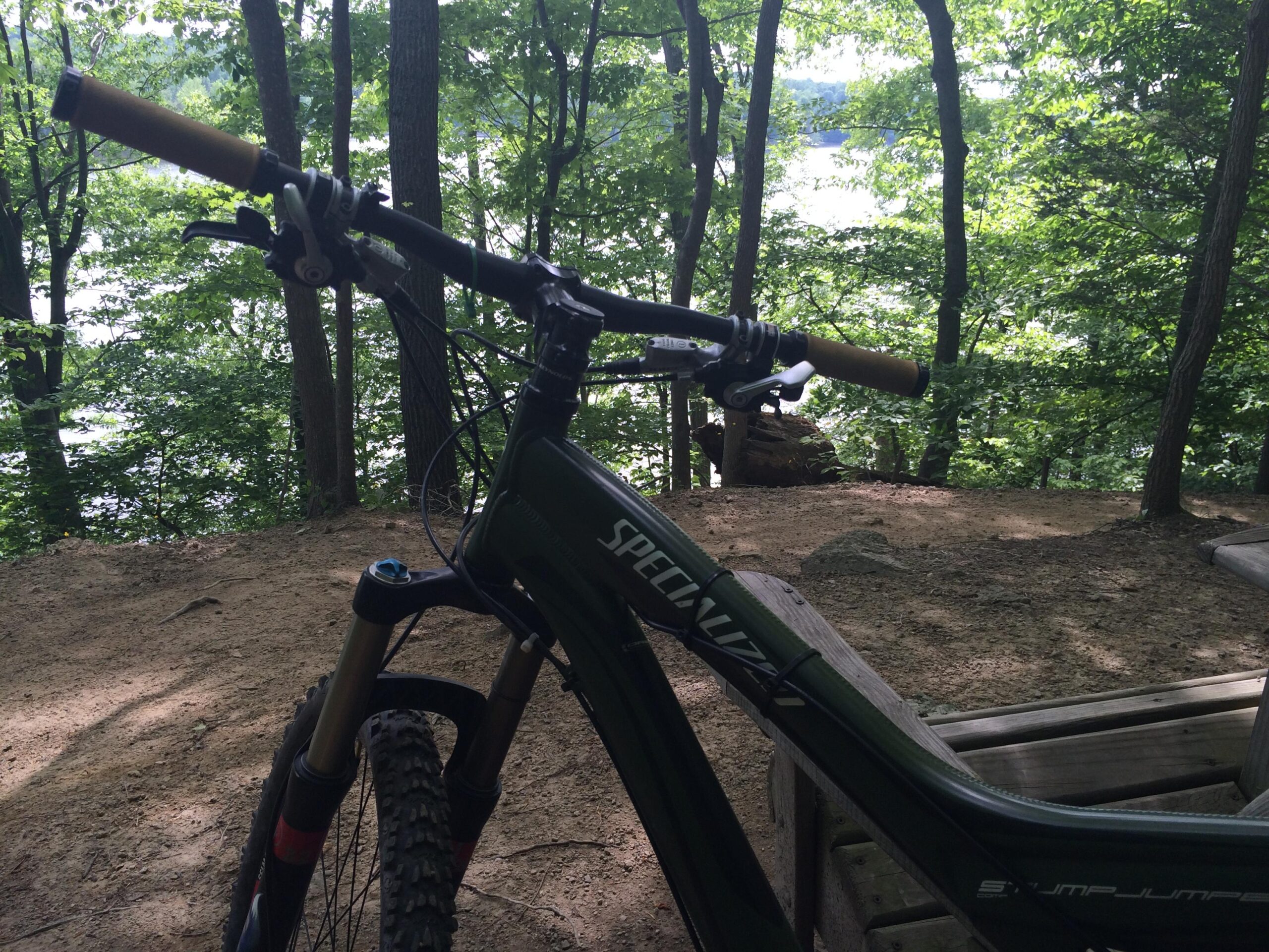 A close-up view of a mountain bike's handlebars with a natural forest backdrop, featuring green trees and a visible body of water in the distance. The bike has brown grips and thick, knobby tires. The ground is sandy and unpaved, suggesting a trail or outdoor setting. Fountainhead Regional Park mountain bike trail.