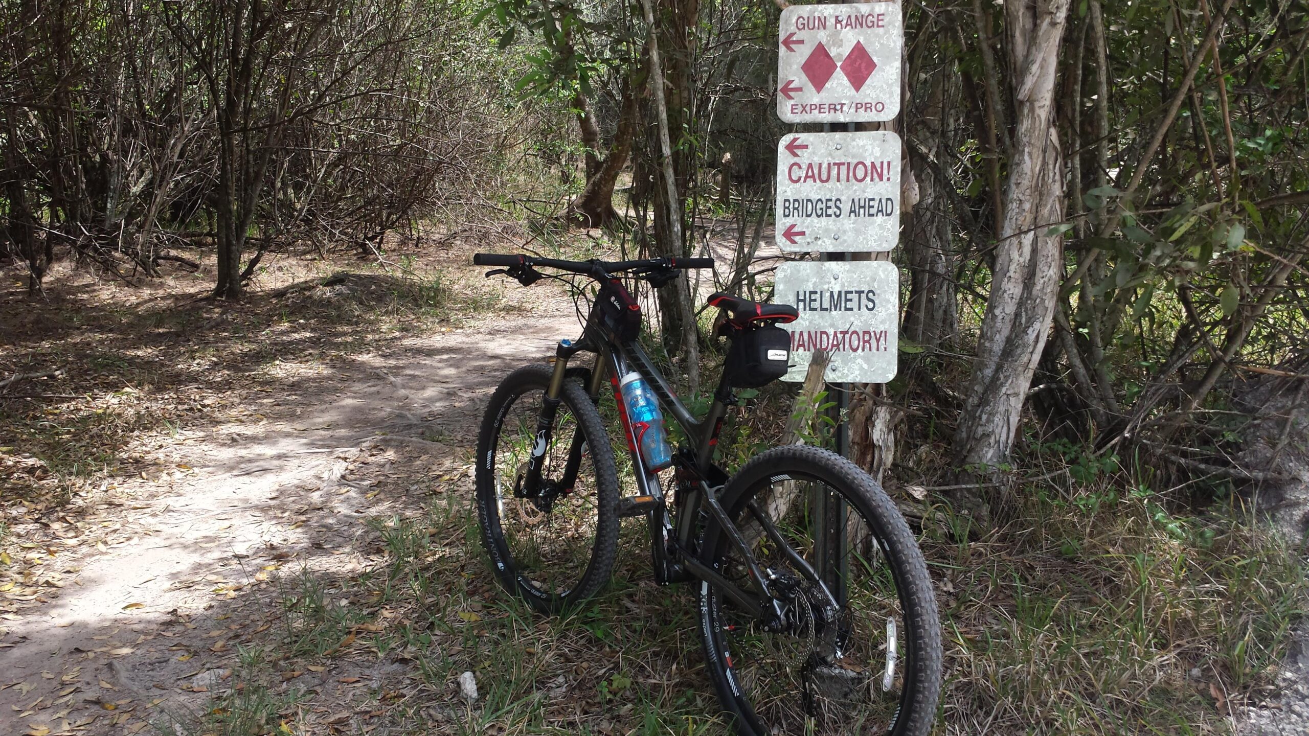 Trek Fuel EX 8: A mountain bike parked on a dirt path surrounded by trees, with several signs nearby indicating a gun range, caution for bridges ahead, and a reminder that helmets are mandatory.