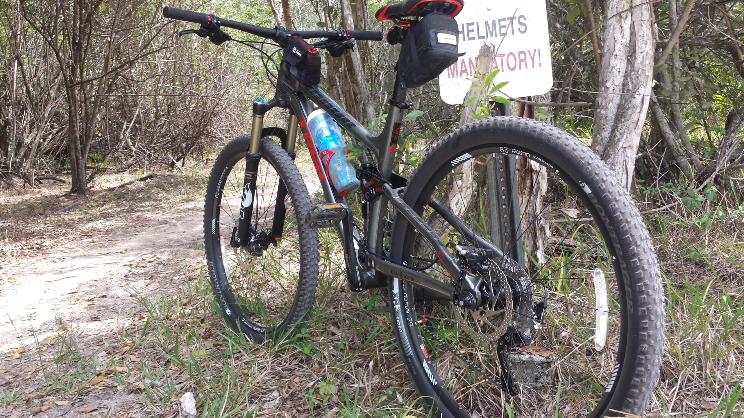 Trek Fuel EX 8: A mountain bike leaning against a sign that reads "HELMETS MANDATORY!" The bike is positioned on a dirt trail surrounded by sparse vegetation and trees. A water bottle is attached to the frame, and the tires are designed for off-road riding.
