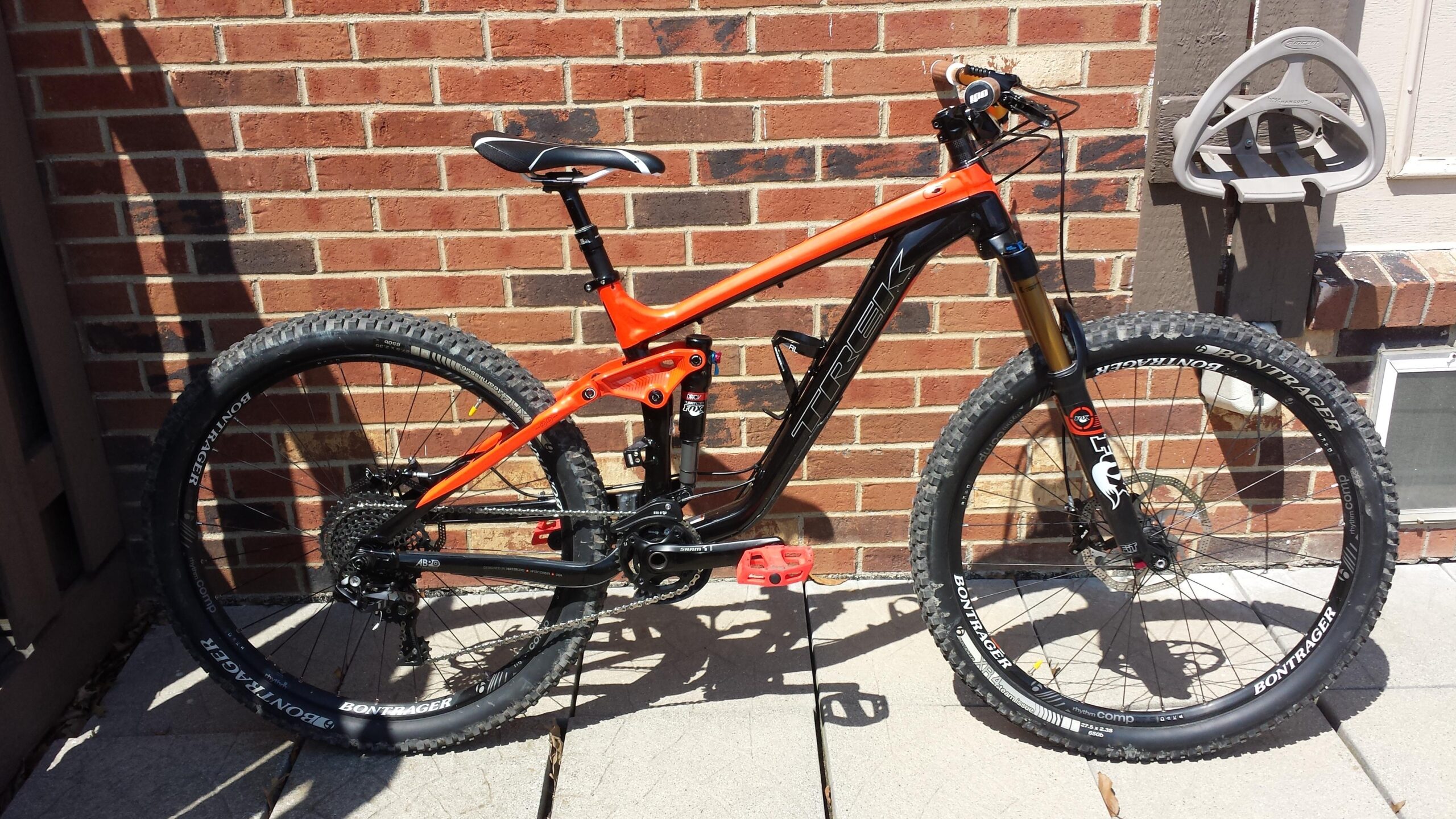 Trek Slash: A close-up image of a Trek mountain bike featuring a black and orange frame. The bike is positioned against a brick wall and sitting on a concrete surface. It has thick, knobby tires and a suspension system, indicating its suitability for off-road riding. There is a bike rack visible in the background.