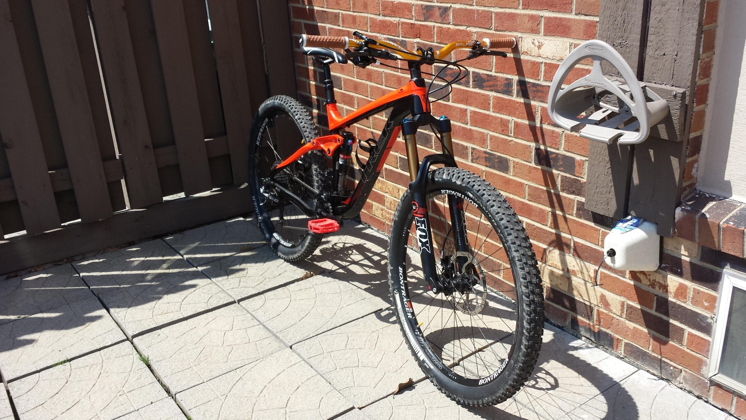 Trek Slash: A bright orange and black mountain bike leans against a brick wall. It features thick, knobby tires and distinctive handlebars with wooden grips. In the background, there is a wooden fence and a bike rack mounted on the wall, alongside a small container. The ground is paved with patterned concrete slabs, and the scene is illuminated by sunlight.