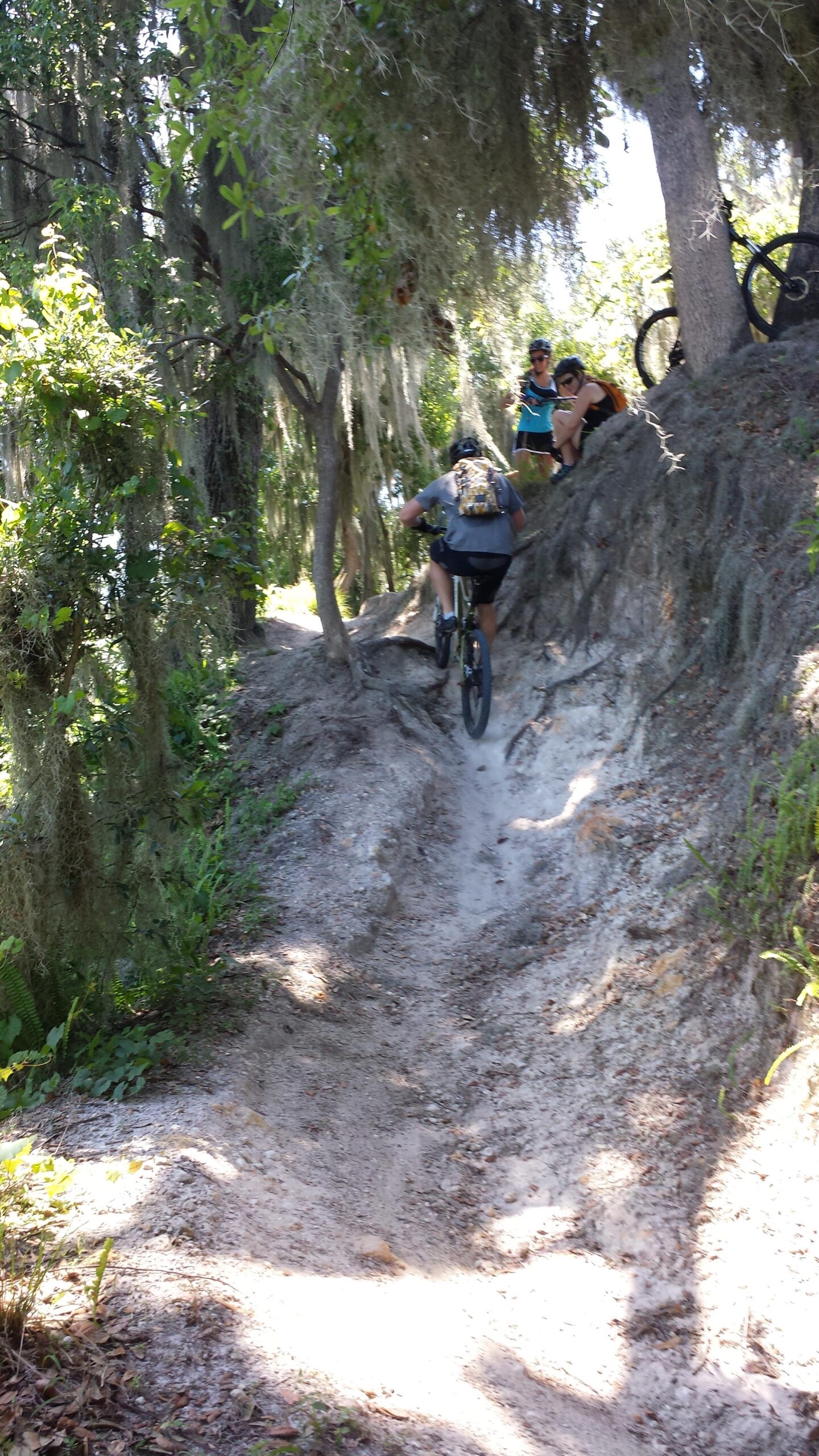 A mountain biker navigating a narrow dirt trail surrounded by trees, with other cyclists observing nearby. Sunlight filters through the foliage, highlighting the sandy path and the lush greenery around. Loyce E. Harpe Park mountain bike trail.