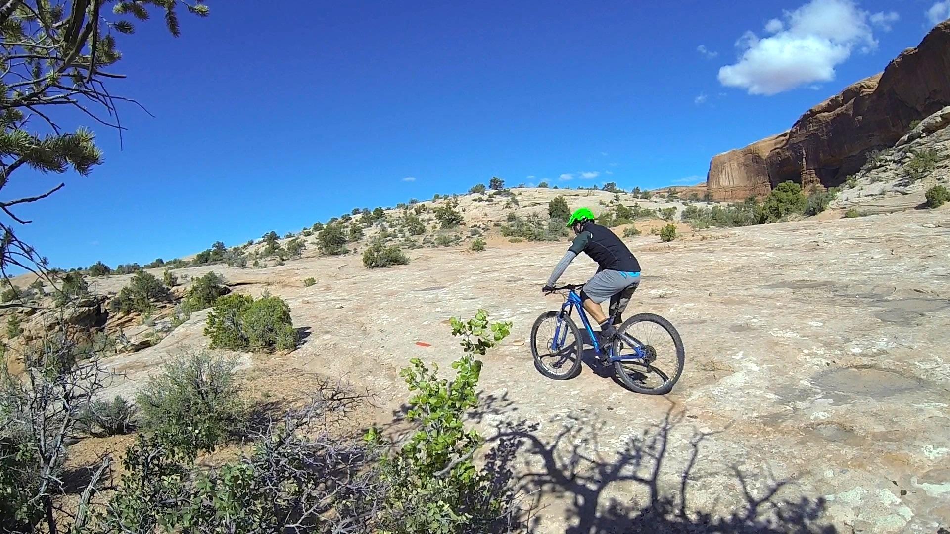 A person riding a blue mountain bike across a rocky landscape under a clear blue sky, with scattered greenery and distant hills in the background. The rider is wearing a black shirt, gray shorts, and a bright green helmet. Ramblin' mountain bike trail.