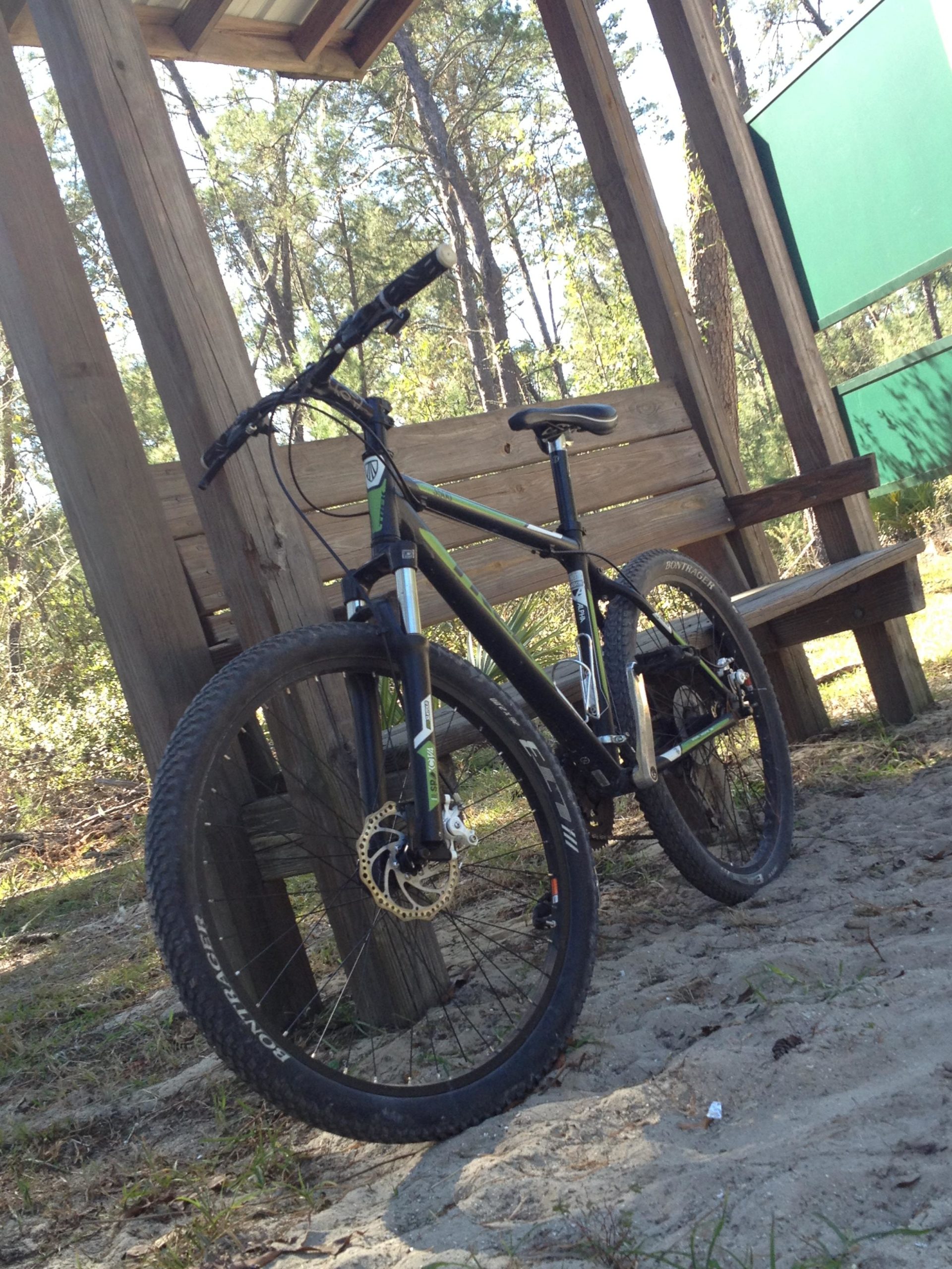A black and green mountain bike is parked beside a wooden bench under a shelter in a wooded area, with trees and brush visible in the background. The ground is sandy and the bike features noticeable dirt and wear, indicating recent use. Santos mountain bike trail.