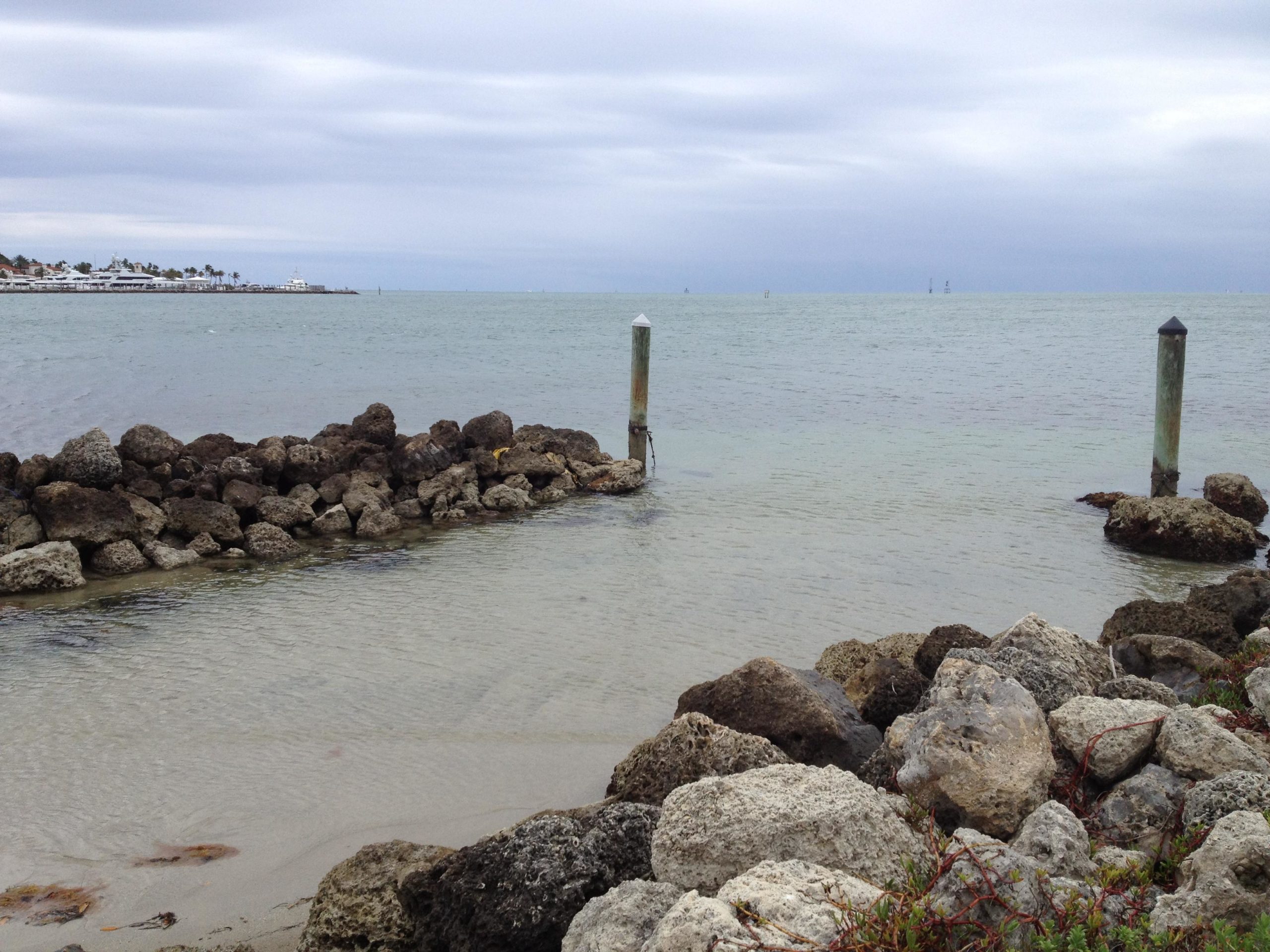 A serene coastal scene featuring smooth waters under a cloudy sky, with rocky shorelines in the foreground. Two weathered posts stand near the water's edge, while distant boats can be seen sailing on the horizon. The landscape evokes a calm and tranquil atmosphere. Virginia Key North Point mountain bike trail.