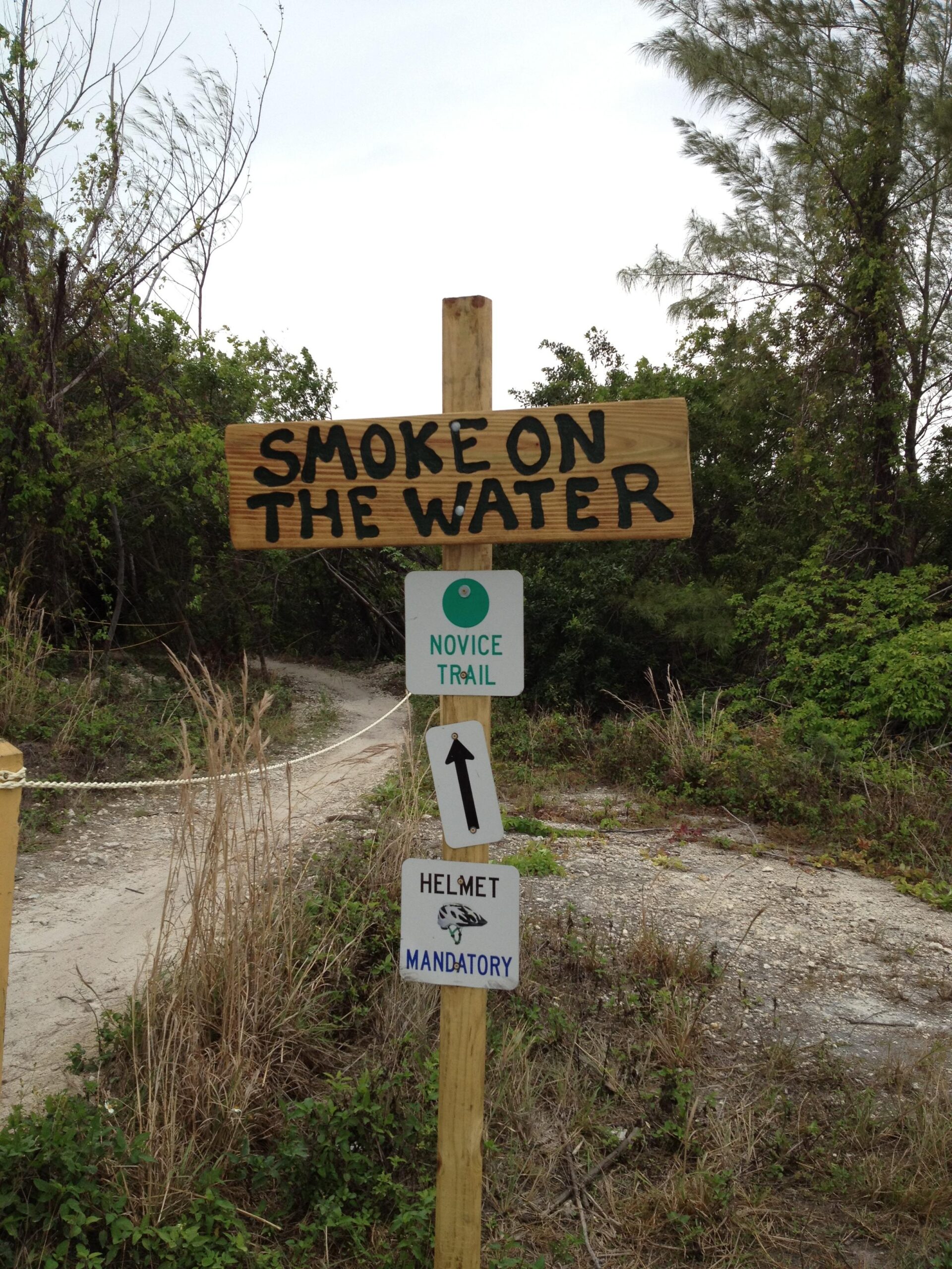 Signpost at the entrance of a novice trail named "Smoke on the Water," indicating that wearing a helmet is mandatory. The trail path is visible in the background, surrounded by greenery. Virginia Key North Point mountain bike trail.
