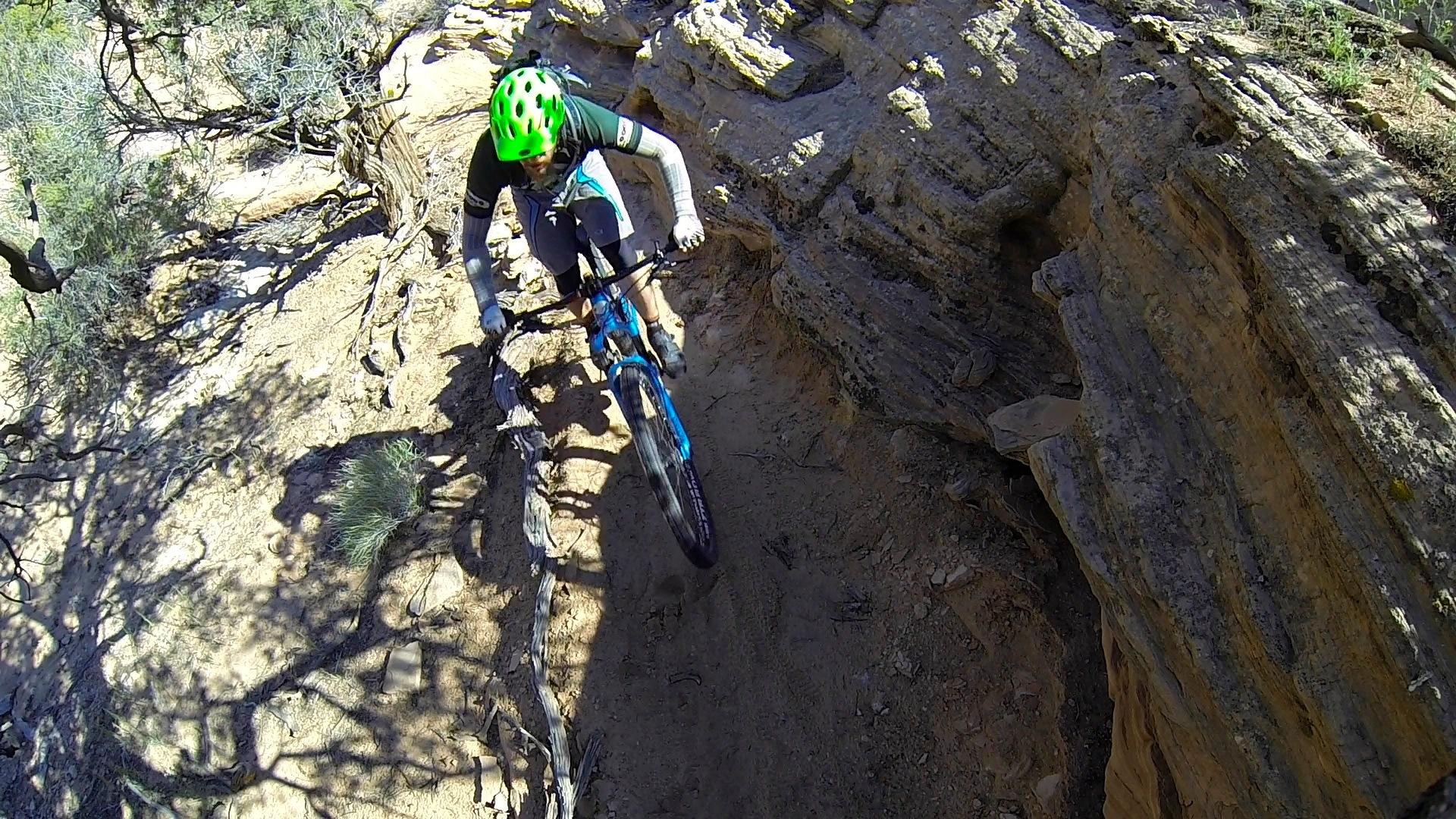 A mountain biker with a green helmet navigates a rocky trail, leaning into a turn on a dirt path surrounded by shrubs and rocky terrain. The scene captures the excitement of off-road biking in a natural landscape. Rocky Tops mountain bike trail.