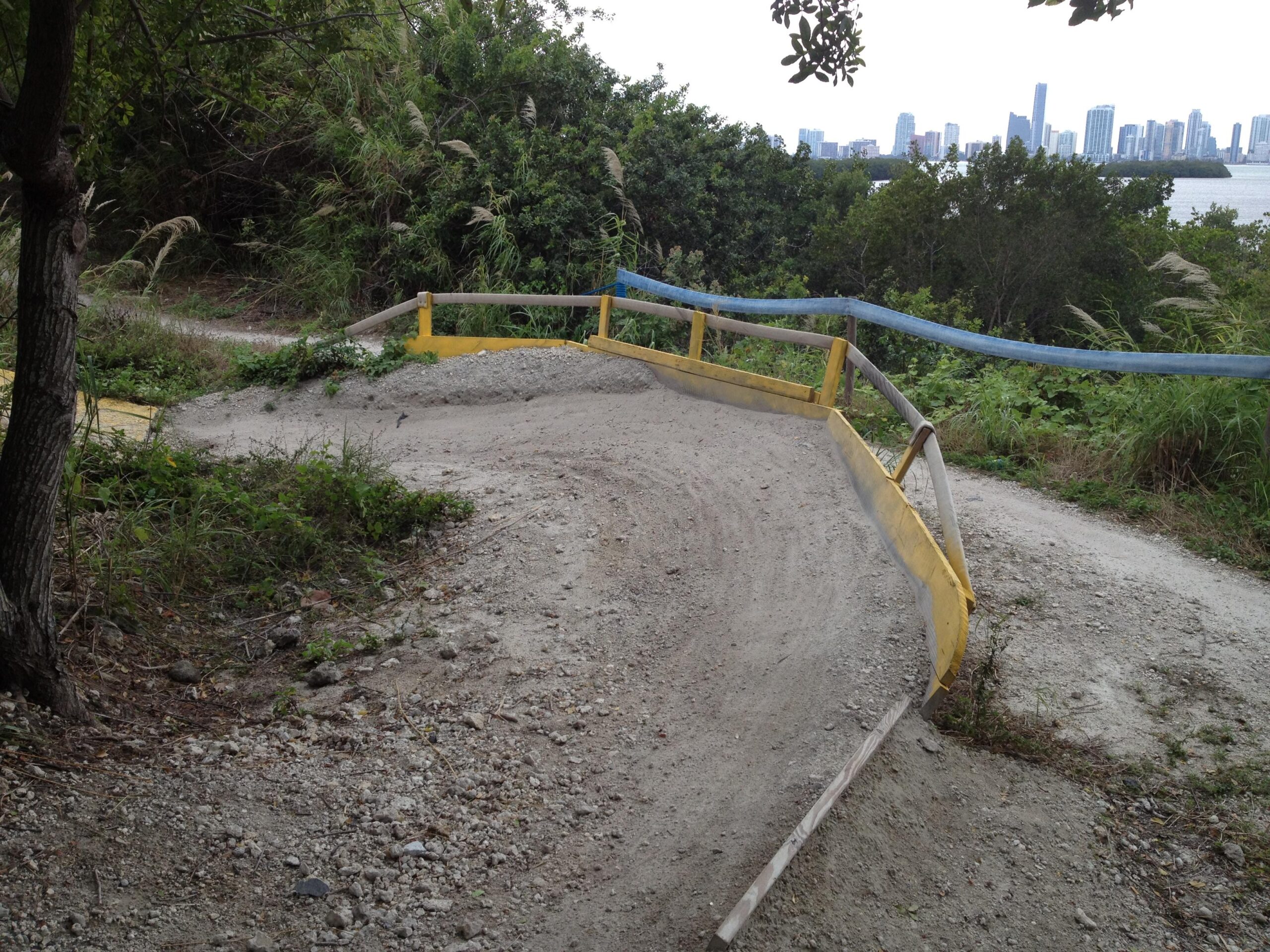 A sandy, winding bike trail with yellow wooden barriers, surrounded by dense greenery and a distant city skyline along a waterfront. Virginia Key North Point mountain bike trail.