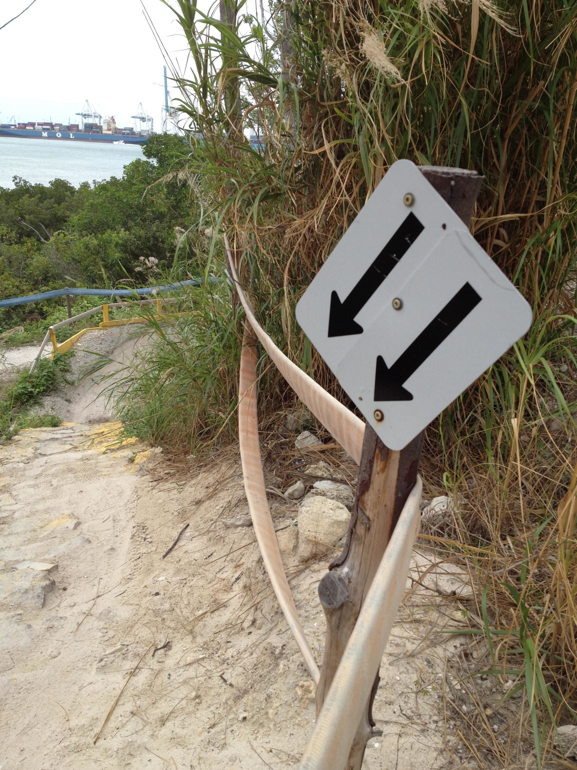 A dirt path leading down towards a body of water, lined with tall grasses and bushes. A directional sign with black arrows pointing up and down is mounted on a wooden post, indicating the trail's direction. In the background, cargo ships can be seen in the water. Virginia Key North Point mountain bike trail.