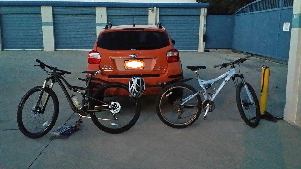 Specialized Rumor Comp: Two mountain bikes are parked next to an orange SUV in a dimly lit parking area. The bikes are positioned at an angle, showcasing their designs, while the SUV is visible in the background alongside storage units. The scene suggests a post-ride setting, with a helmet resting on the bike's handlebars.