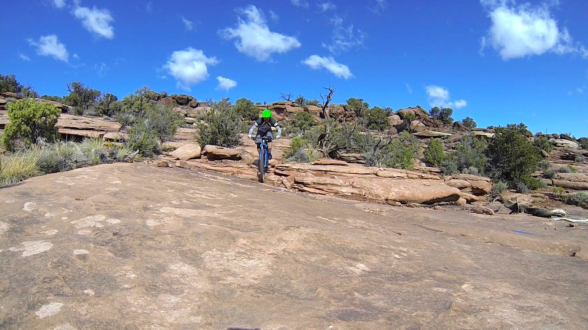 A mountain biker navigates a rocky trail under a clear blue sky dotted with fluffy clouds. The landscape features rugged terrain with various shrubs and trees in the background. Captain Ahab mountain bike trail.