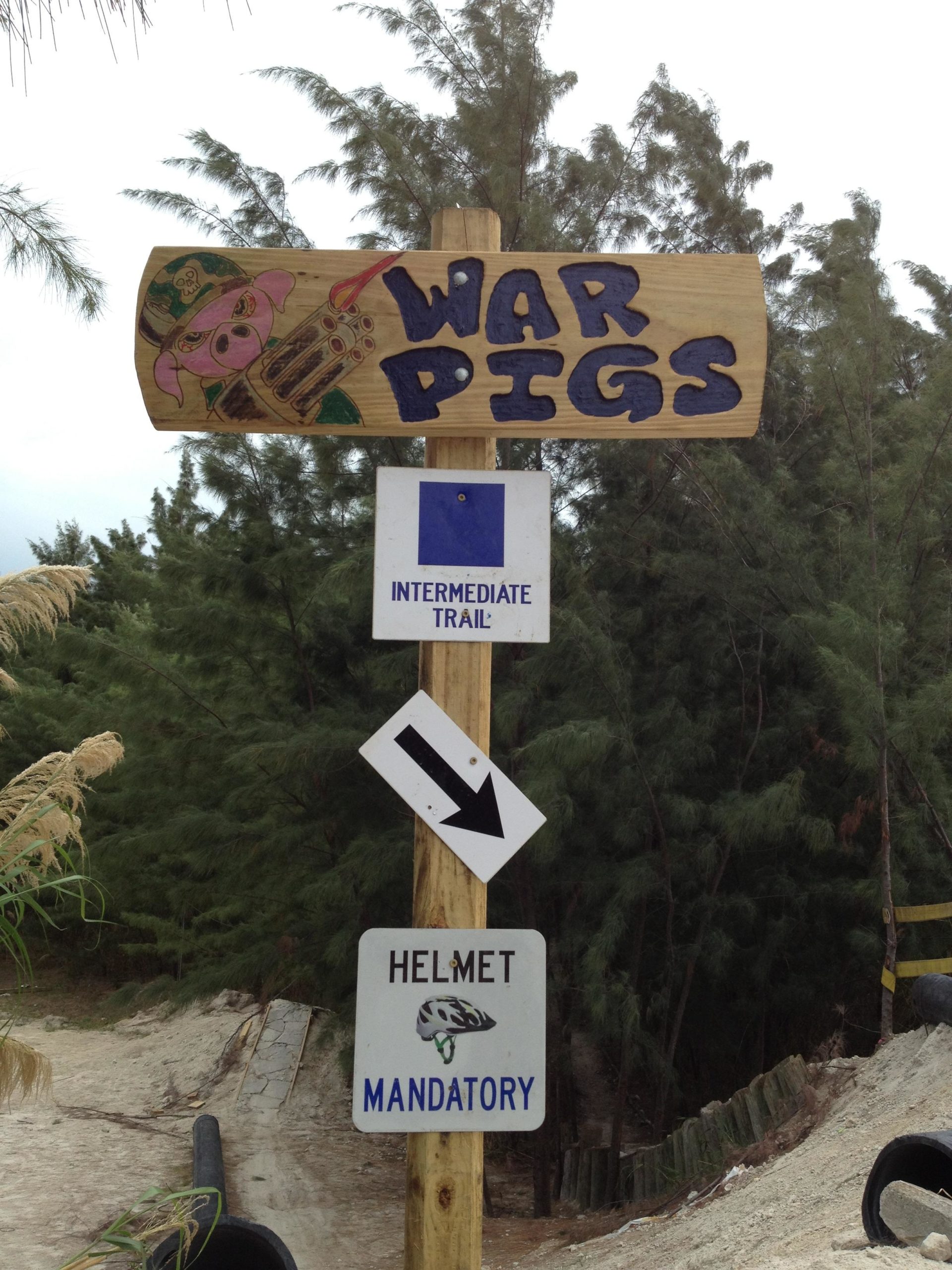 A wooden signpost with the title "WAR PIGS" at the top, featuring a cartoon pig character with a weapon. Below, there is a sign indicating an "INTERMEDIATE TRAIL" with a blue square symbol. An arrow points downward to another sign stating "HELMET MANDATORY" along with a graphic of a helmet. In the background, there are green trees and a sandy path. Virginia Key North Point mountain bike trail.