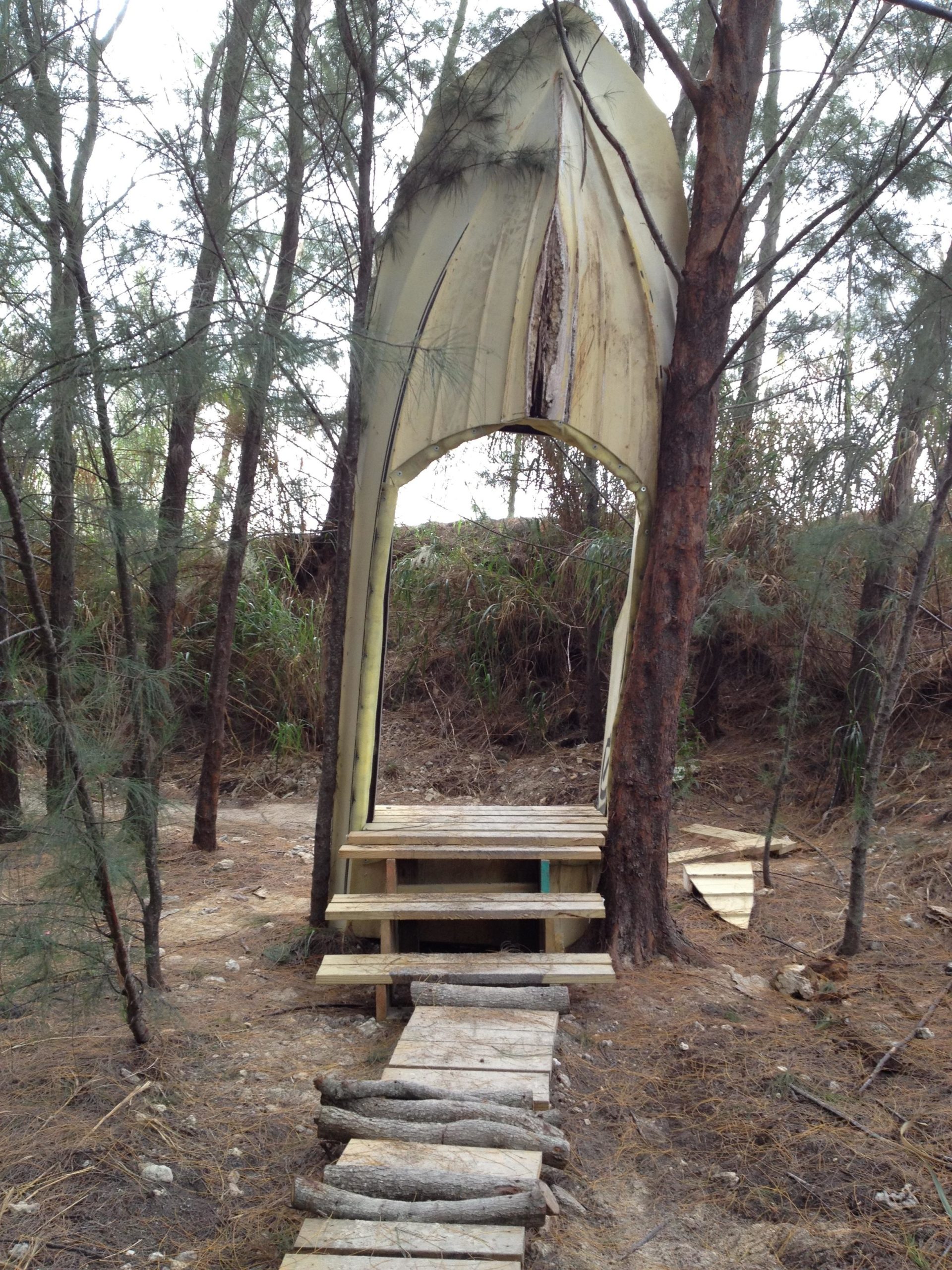 A wooden pathway leads up to a large, archway structure resembling an open door, situated in a forested area. The archway is bordered by tall trees and features a distinct, pointed top. Surrounding vegetation is sparse, with some underbrush visible. The structure appears handmade, composed of wood and possibly repurposed materials. Virginia Key North Point mountain bike trail.