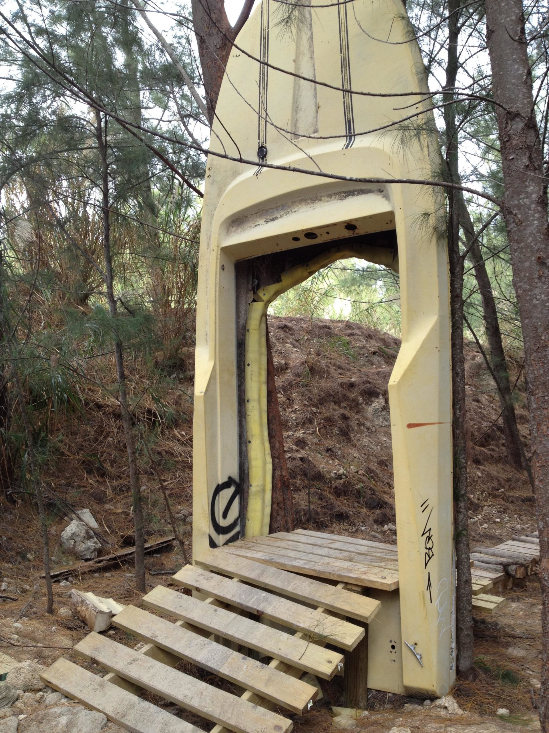 An unusual entryway resembling the shape of a large surfboard is partially embedded in the forest. It features a yellow and white color scheme with some graffiti markings on it. A wooden path made of planks leads up to it, surrounded by trees and natural foliage. Virginia Key North Point mountain bike trail.