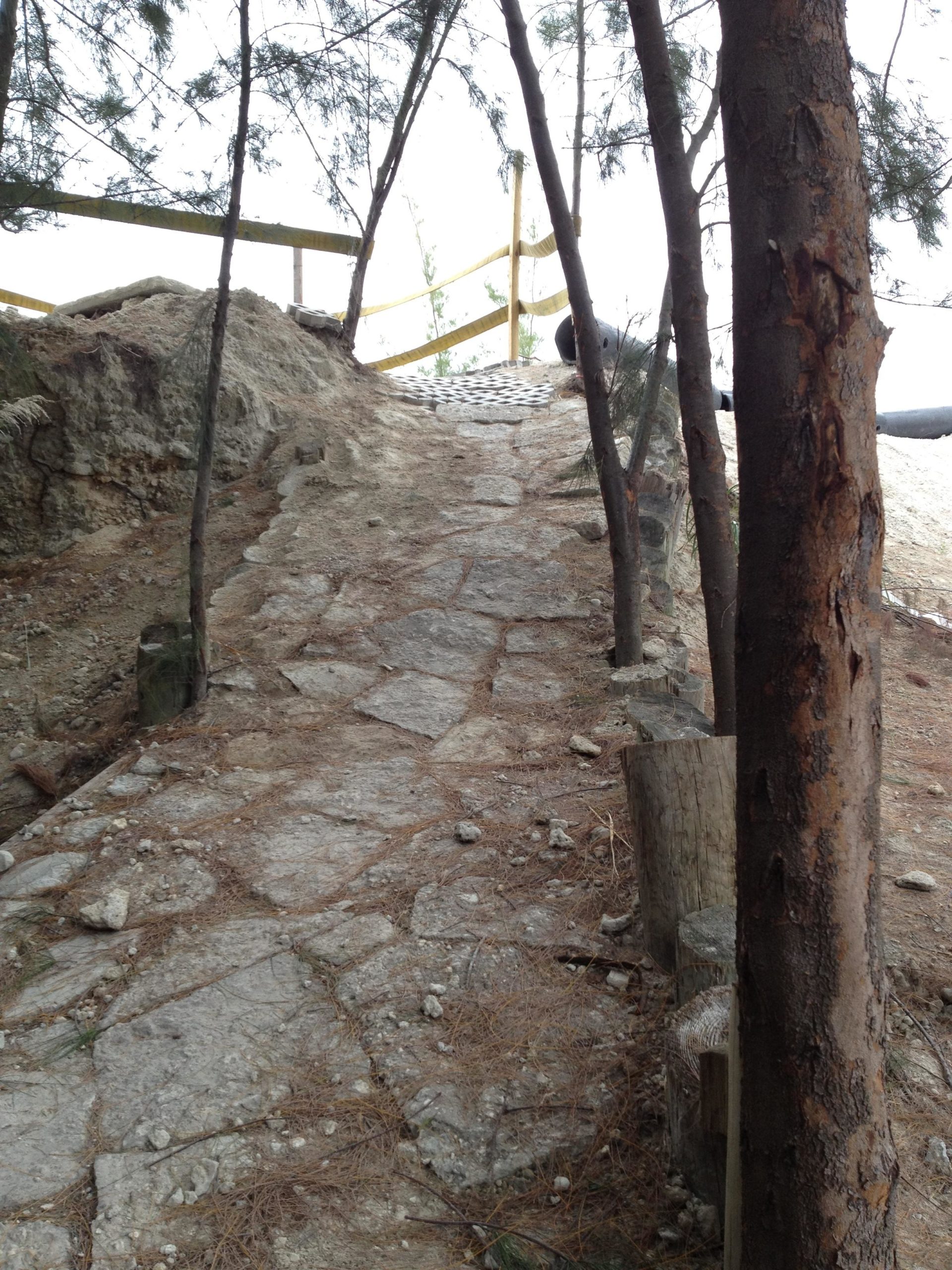 A narrow stone pathway leading up a slope, surrounded by trees and natural terrain. A wooden railing is partially visible in the background, providing a boundary along the path. The ground is covered with pine needles and small stones, creating a rustic outdoor scene. Virginia Key North Point mountain bike trail.