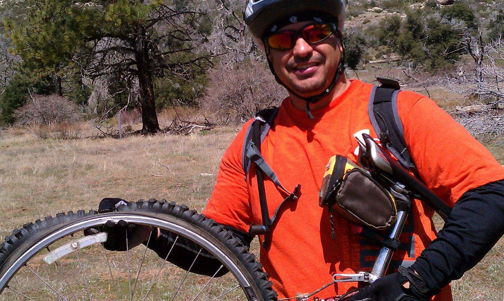 Trek 930: A person wearing a helmet and sunglasses holds a mountain bike tire while standing in a grassy area with trees in the background. The individual is dressed in a bright orange shirt and black gloves, showcasing outdoor biking gear.