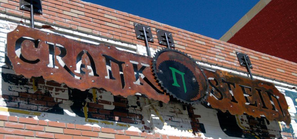 Signage for "Crank n Stein" mounted on a brick wall, featuring a rusted metal design and a circular saw blade graphic integrated into the text. The background shows a clear blue sky and hints of peeling paint on the wall.