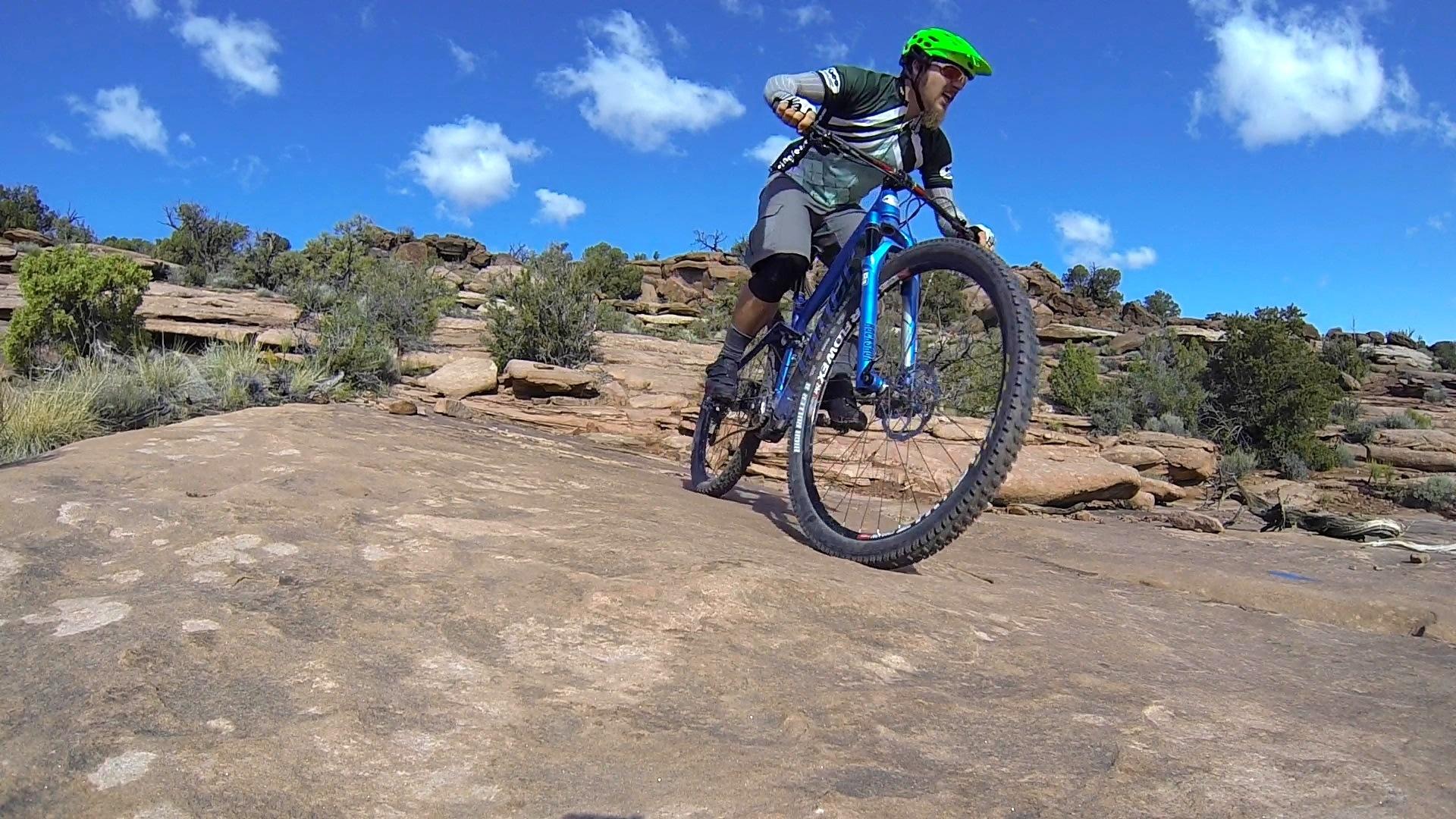A person riding a blue mountain bike on rocky terrain, wearing a green helmet and sunglasses. The background features a vibrant blue sky with scattered clouds and shrubs on the rocky landscape. The rider is leaning forward, navigating the uneven surface. Captain Ahab mountain bike trail.