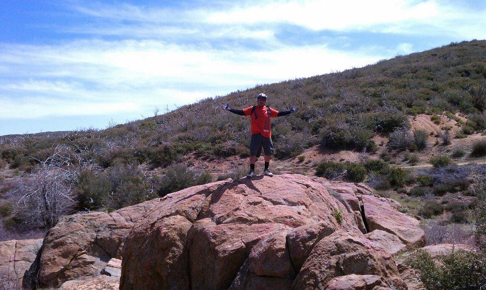 A person stands on top of a large rock, arms outstretched in a pose of triumph, against a backdrop of rolling hills and a clear blue sky. The landscape features sparse vegetation and rocky terrain, suggesting an outdoor hiking or biking adventure. The individual is wearing an orange shirt and shorts, along with a helmet. Cuyamaca Rancho State Park mountain bike trail.