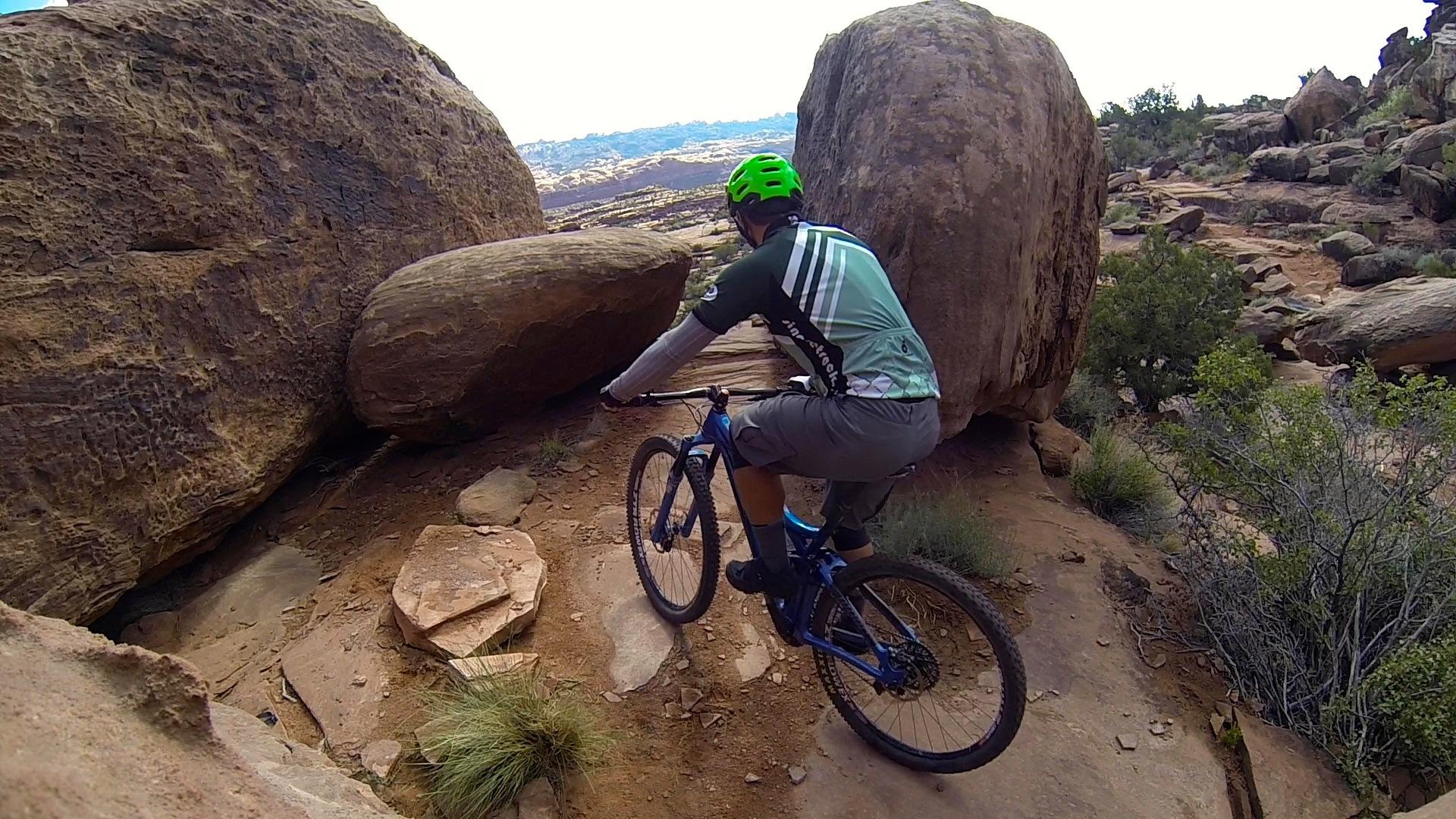 A mountain biker in a green helmet navigates a rocky trail between large boulders, surrounded by sparse vegetation and a hilly landscape in the background. Captain Ahab mountain bike trail.