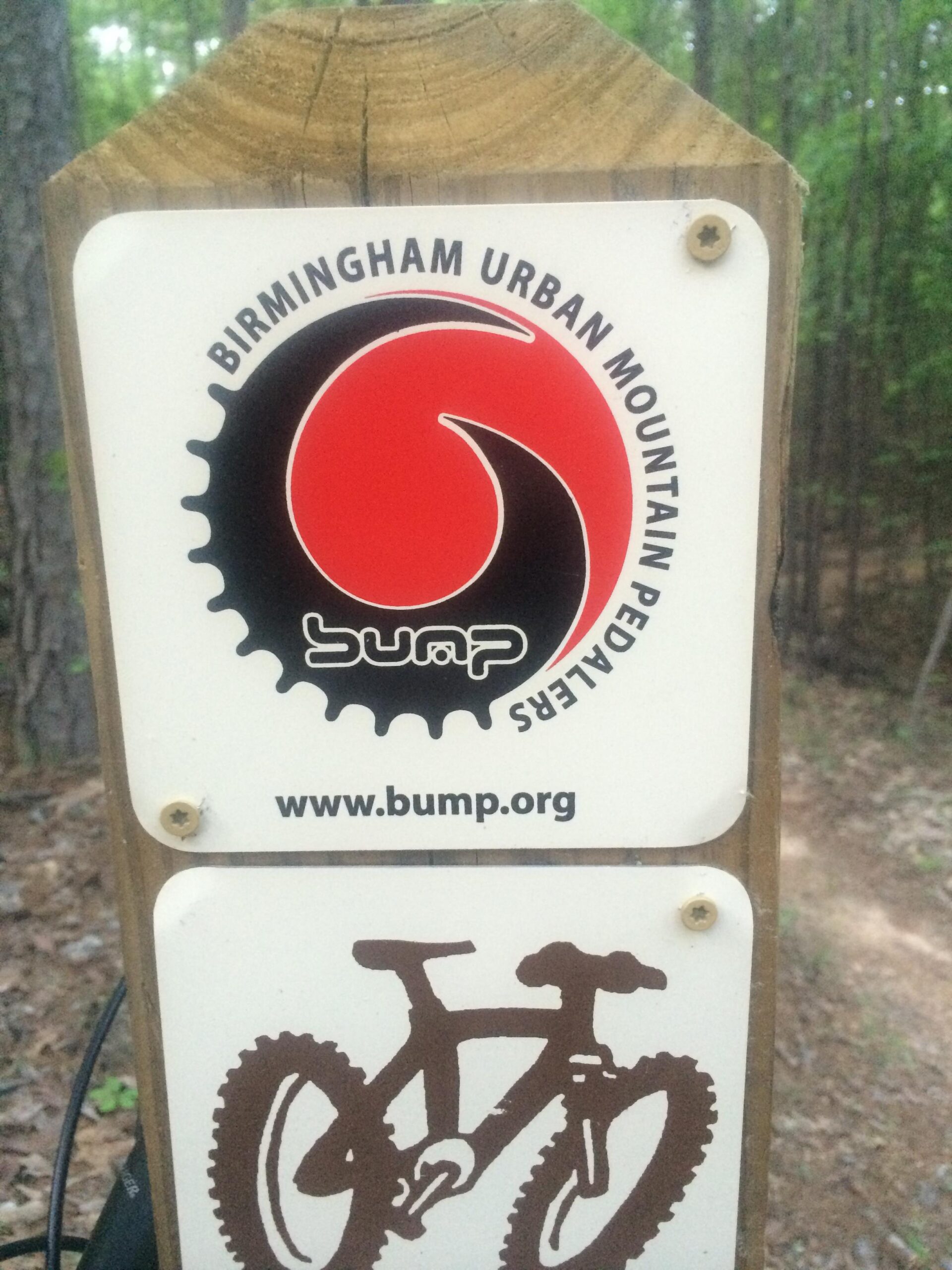 Sign for Birmingham Urban Mountain Pedalers (BUMP) featuring a logo with a stylized red swirl and a bicycle graphic. The sign includes the website URL "www.bump.org" and is mounted on a wooden post, set against a forested background.