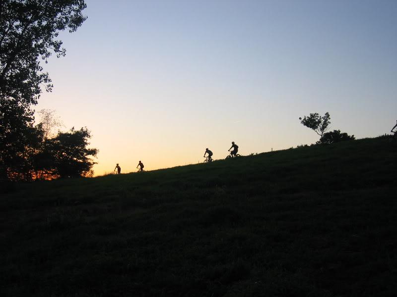 Silhouettes of five cyclists riding up a grassy hill during sunset, with trees framing the scene against a colorful sky.