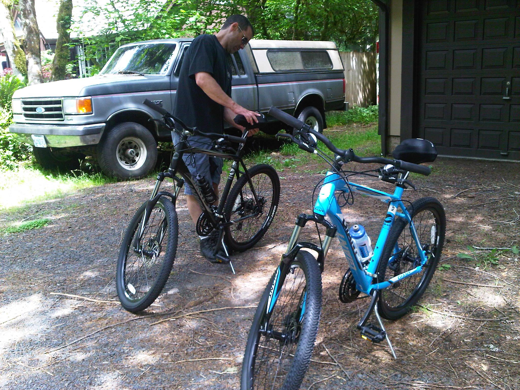 Giant Revel 0: A man wearing a black t-shirt and sunglasses is standing next to two mountain bikes, one black and one blue, in a gravel area surrounded by trees. A gray pickup truck is parked in the background.