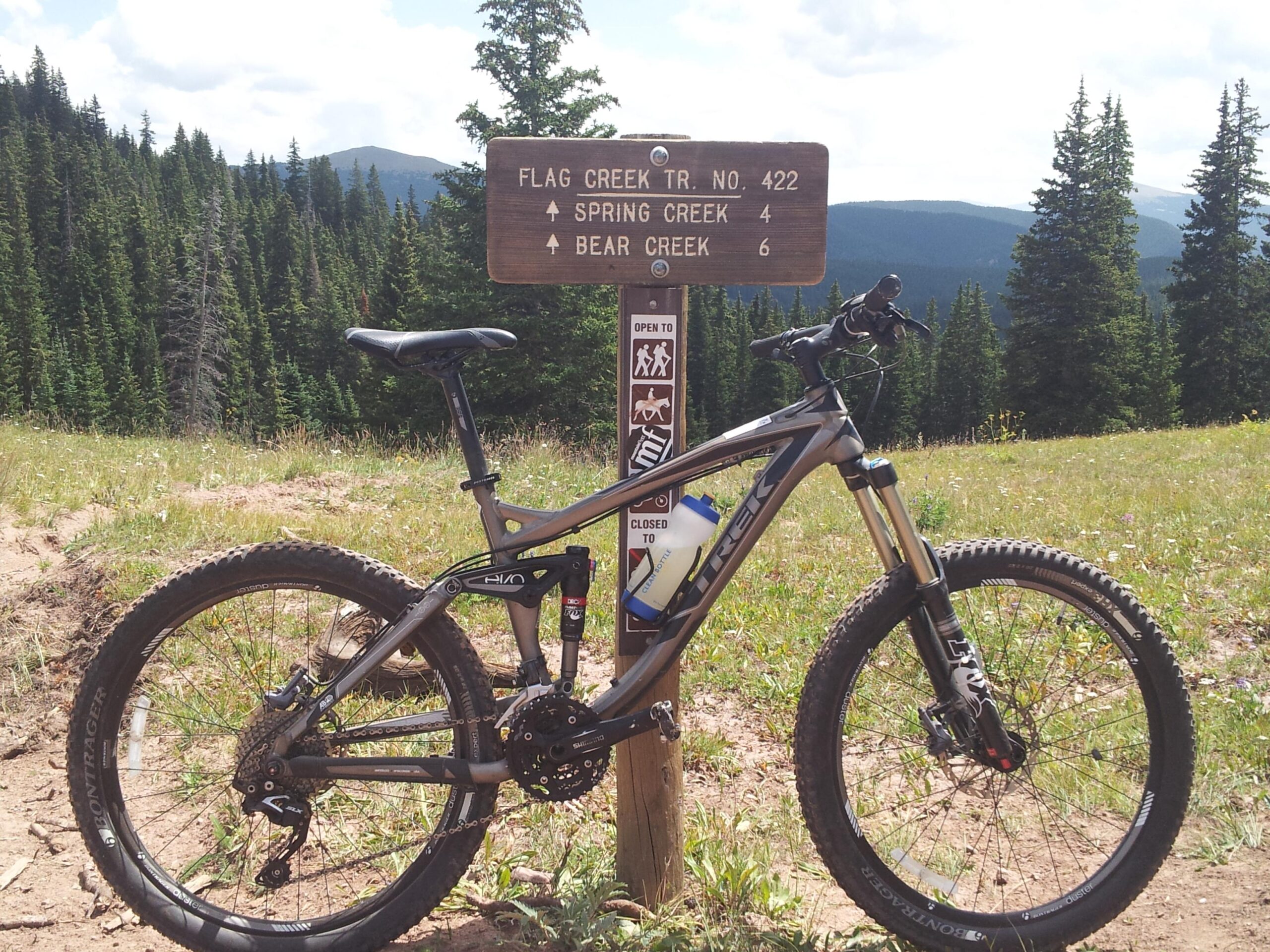 Trek Remedy 7: A mountain bike parked next to a trail sign indicating "Flag Creek Trail No. 422," with distances to "Spring Creek 4 miles" and "Bear Creek 6 miles," set against a scenic backdrop of evergreen trees and mountains under a partly cloudy sky.