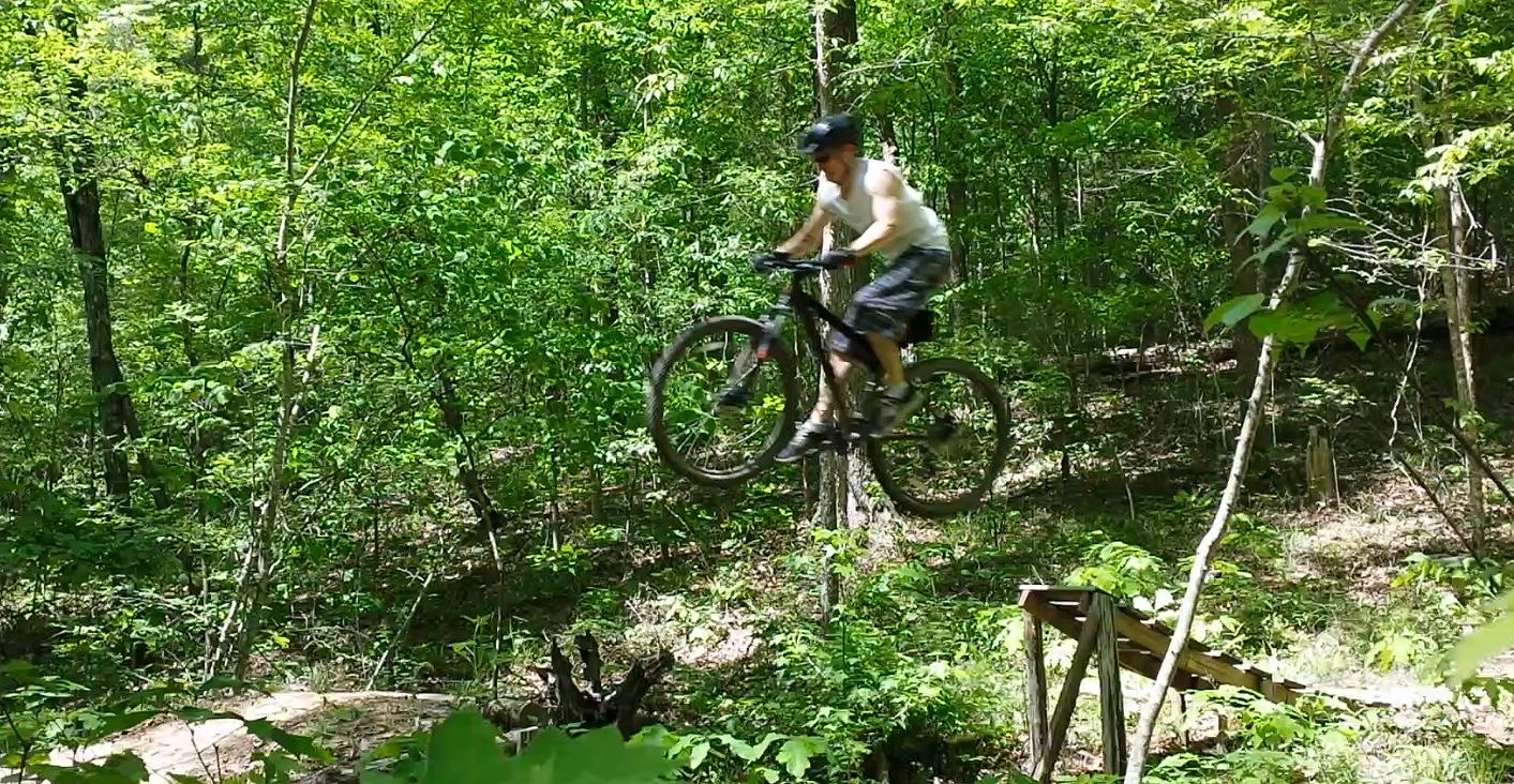 Specialized Hardrock Sport Disc 29er: A person riding a mountain bike mid-air while jumping off a wooden ramp in a lush green forest.