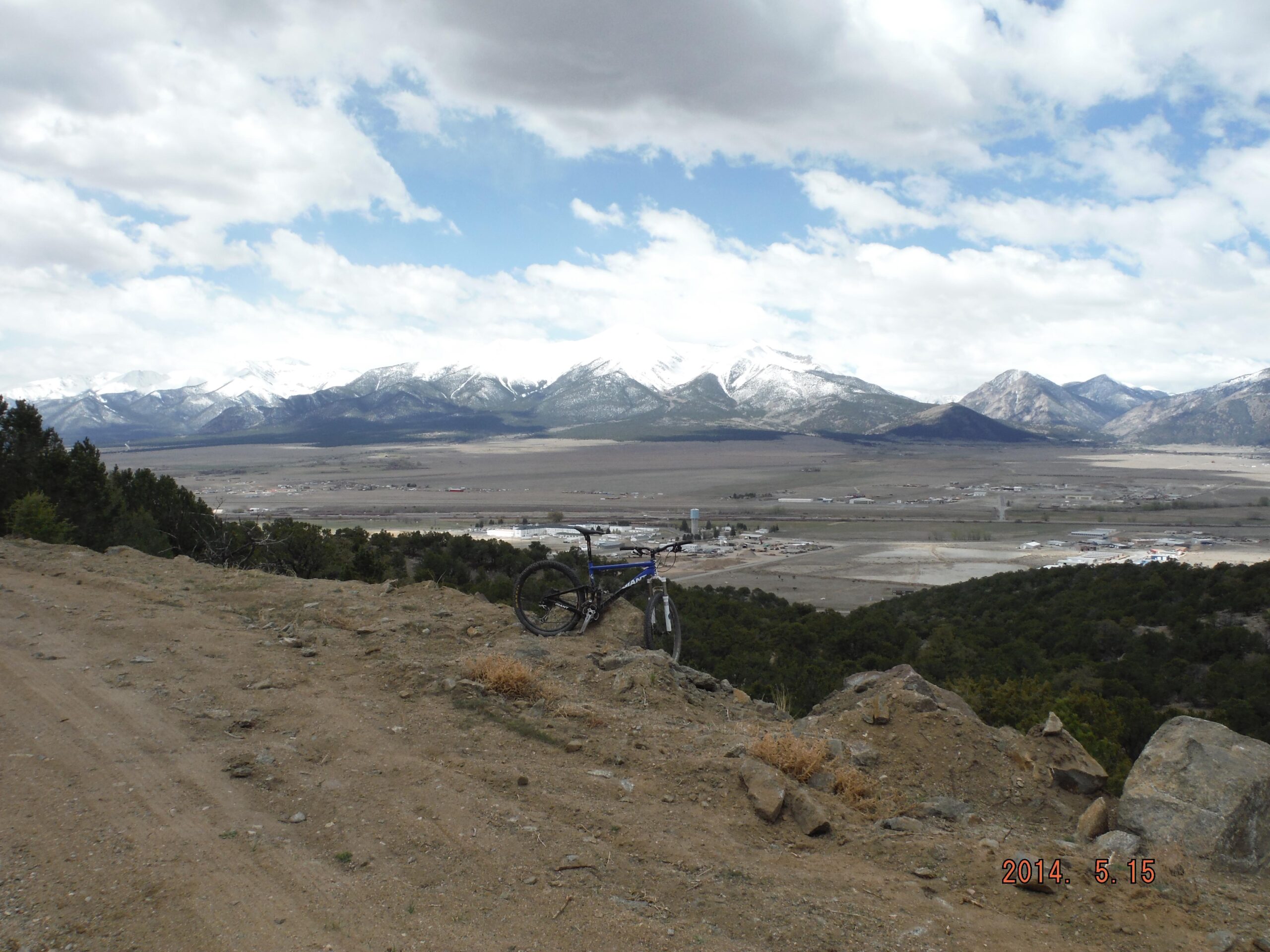 Giant Anthem Advanced: A mountain bike resting on rocky terrain, overlooking a vast valley with a small town, surrounded by snow-capped mountains under a partly cloudy sky.