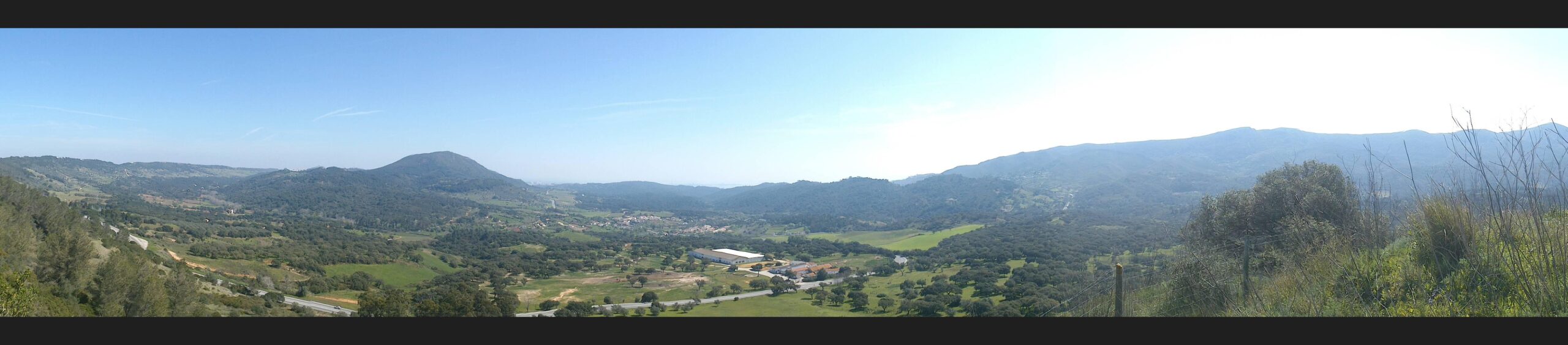 A panoramic view of rolling hills and valleys under a clear blue sky, showcasing lush greenery, scattered trees, and a small village nestled in the landscape. Wisps of clouds are visible in the distance, adding depth to the scenic vista. Arrabida mountain bike trail.