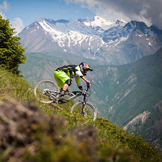 Specialized Enduro Evo: A mountain biker in vibrant green shorts navigates a steep, grassy slope with a backdrop of snow-capped mountains and blue skies. The biker is focused and lean, showcasing dynamic movement and skill.