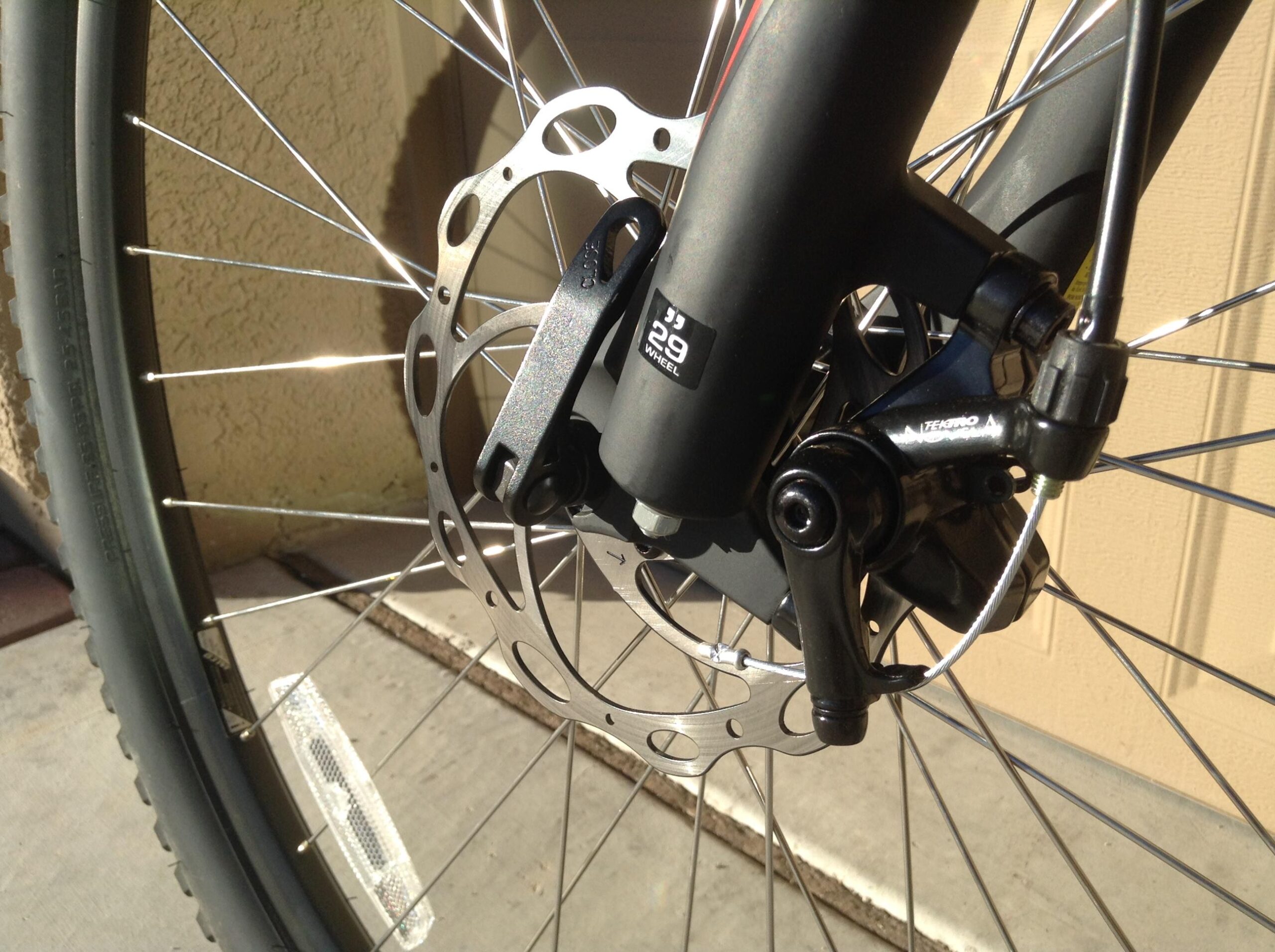 Specialized Hardrock Disc 29: Close-up view of a bicycle's front wheel assembly, showcasing the disc brake system, including the brake rotor and caliper. The image captures the tire, spokes, and part of the bike frame, with a textured background of an exterior wall. Sunlight highlights the metallic components, emphasizing their details.