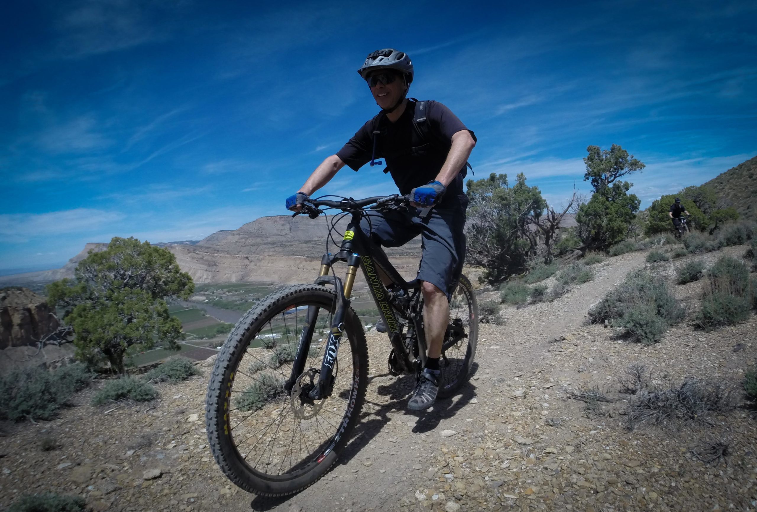A person riding a mountain bike on a dirt trail with rocky terrain, surrounded by sparse vegetation and a mountainous landscape under a blue sky. The cyclist is wearing a helmet and gloves, focused on the ride, with another biker visible in the background. Palisade Rim mountain bike trail.