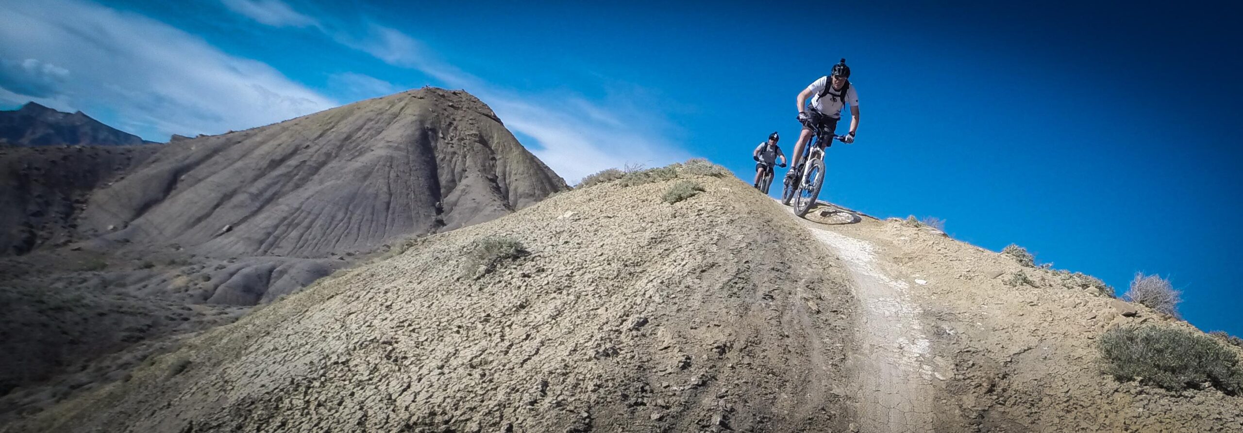 Two mountain bikers riding on a narrow dirt trail along a ridge in a mountainous landscape against a bright blue sky. The terrain is rugged with steep slopes and sparse vegetation. Zippety Do Dah mountain bike trail.