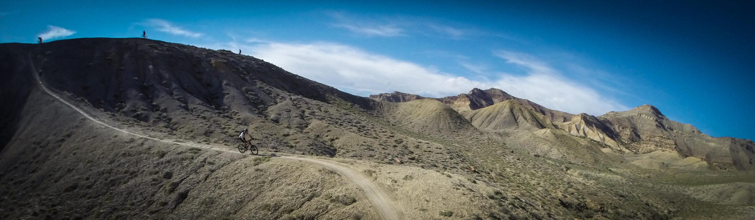 A scenic view of rugged mountains and hills under a bright blue sky, with a winding dirt trail. A cyclist is riding along the trail in the foreground, while other bikers are visible on the ridge in the background. The landscape is arid, featuring sparse vegetation and textured hills. Zippety Do Dah mountain bike trail.