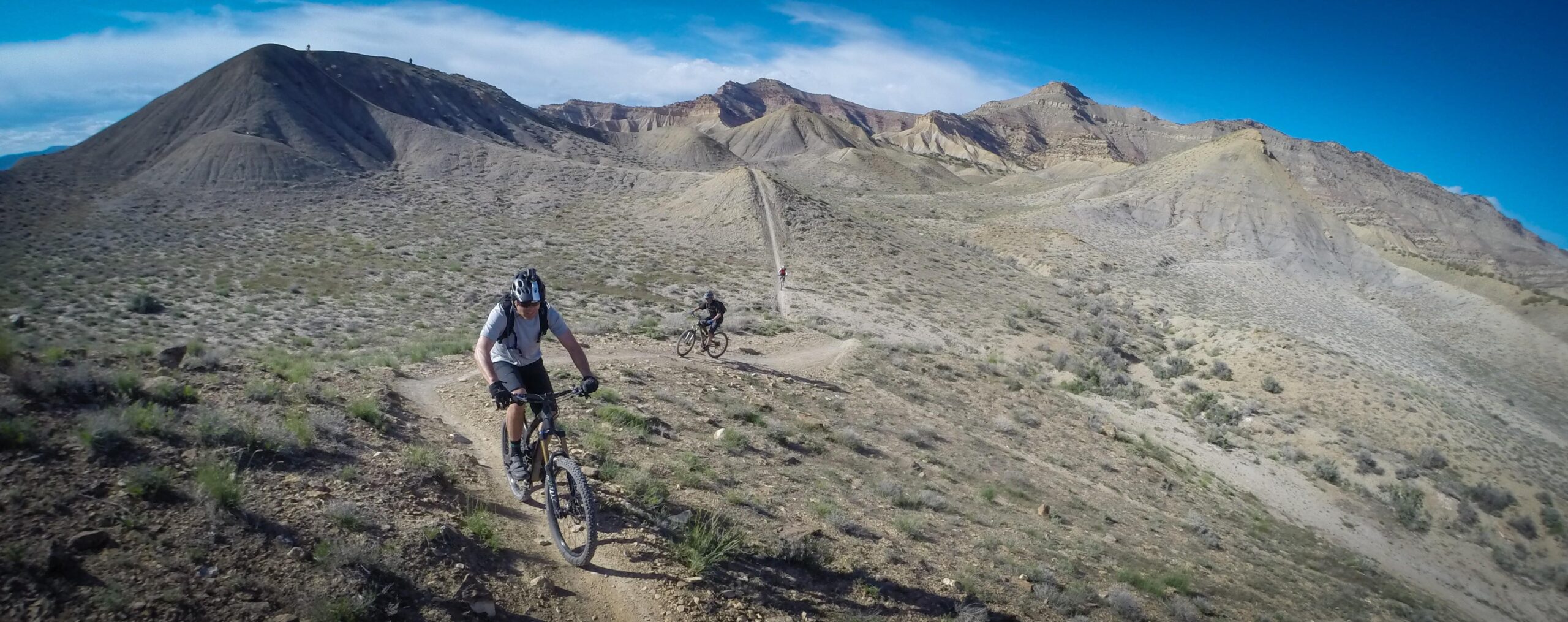 Two mountain bikers navigate a winding dirt trail in a rugged desert landscape, surrounded by hills and rocky terrain under a blue sky. Zippety Do Dah mountain bike trail.