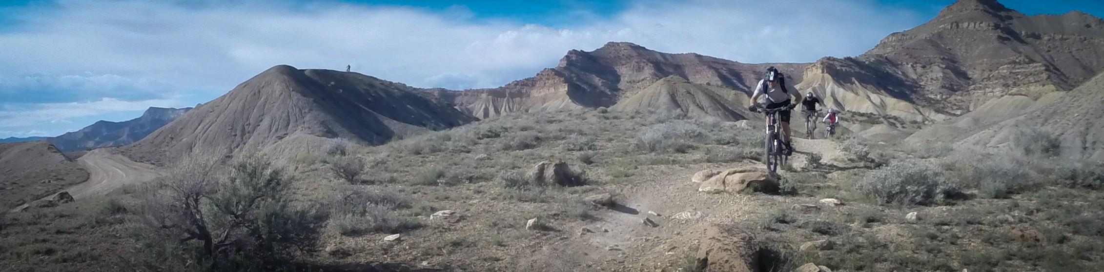 Mountain bikers riding along a dirt trail in a rugged, hilly landscape with sparse vegetation and distant mountains under a partly cloudy sky. Zippety Do Dah mountain bike trail.