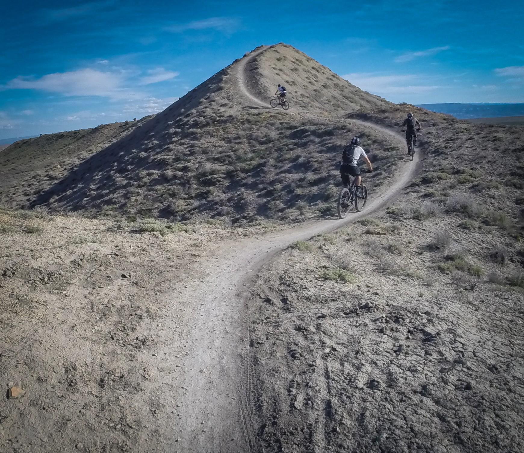 Three mountain bikers navigate a winding dirt trail on a hilly landscape under a bright blue sky. The trail winds up and around a small peak, with sparse vegetation on the hillsides and a clear view of the horizon in the background. Zippety Do Dah mountain bike trail.