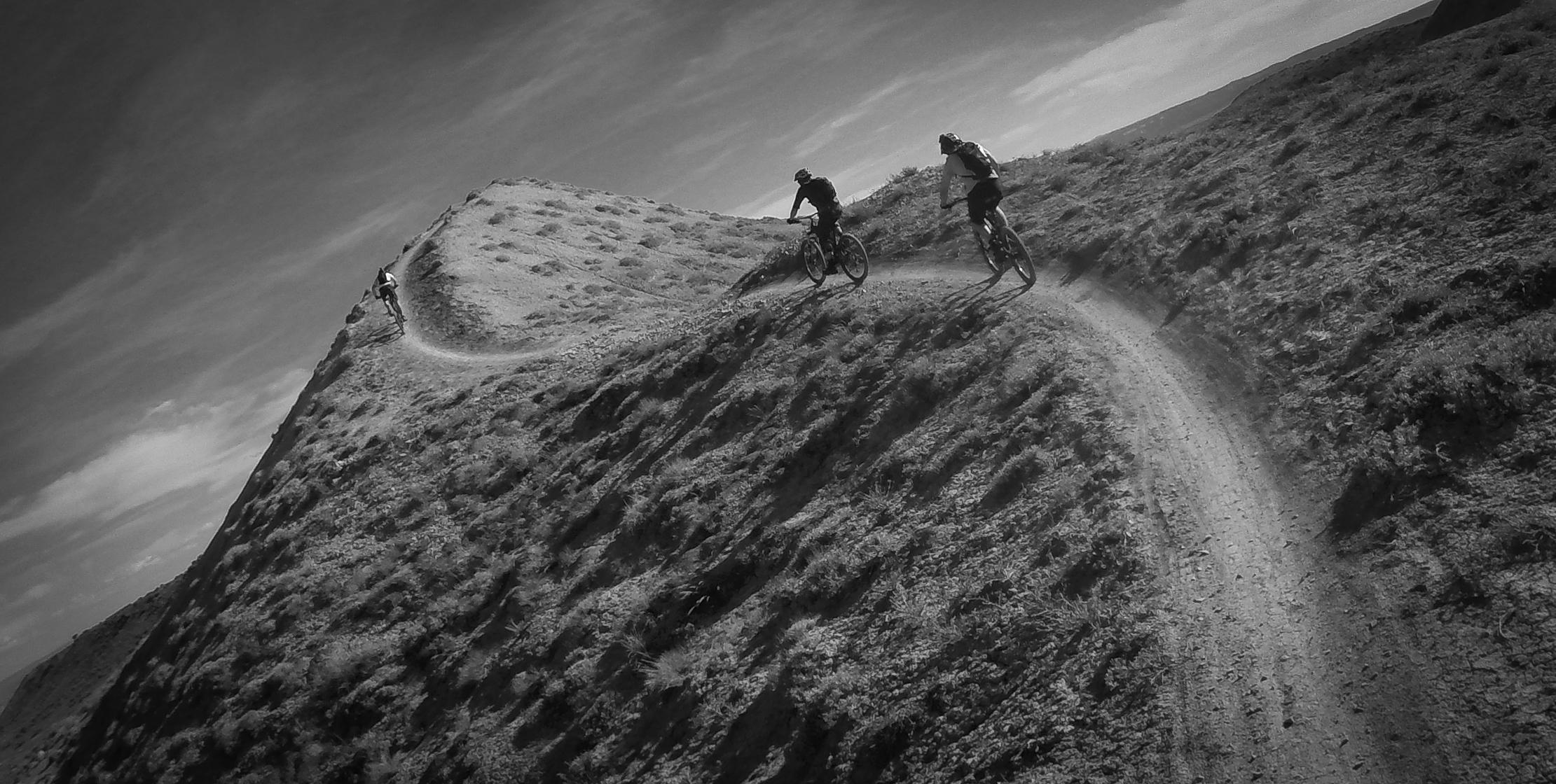Three mountain bikers navigate a winding, rugged dirt trail along a hillside, surrounded by sparse vegetation under a cloudy sky. The image is presented in black and white, highlighting the contours of the landscape and the dynamic motion of the cyclists. Zippety Do Dah mountain bike trail.