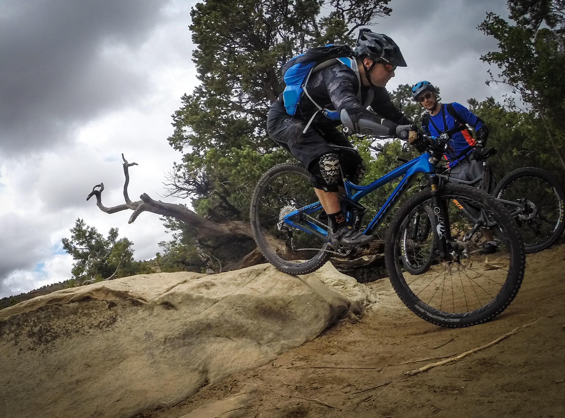 A mountain biker dressed in protective gear jumps off a rocky ledge while another biker looks on. The scene is set in a rugged outdoor environment with trees in the background and a cloudy sky. Porcupine Rim mountain bike trail.